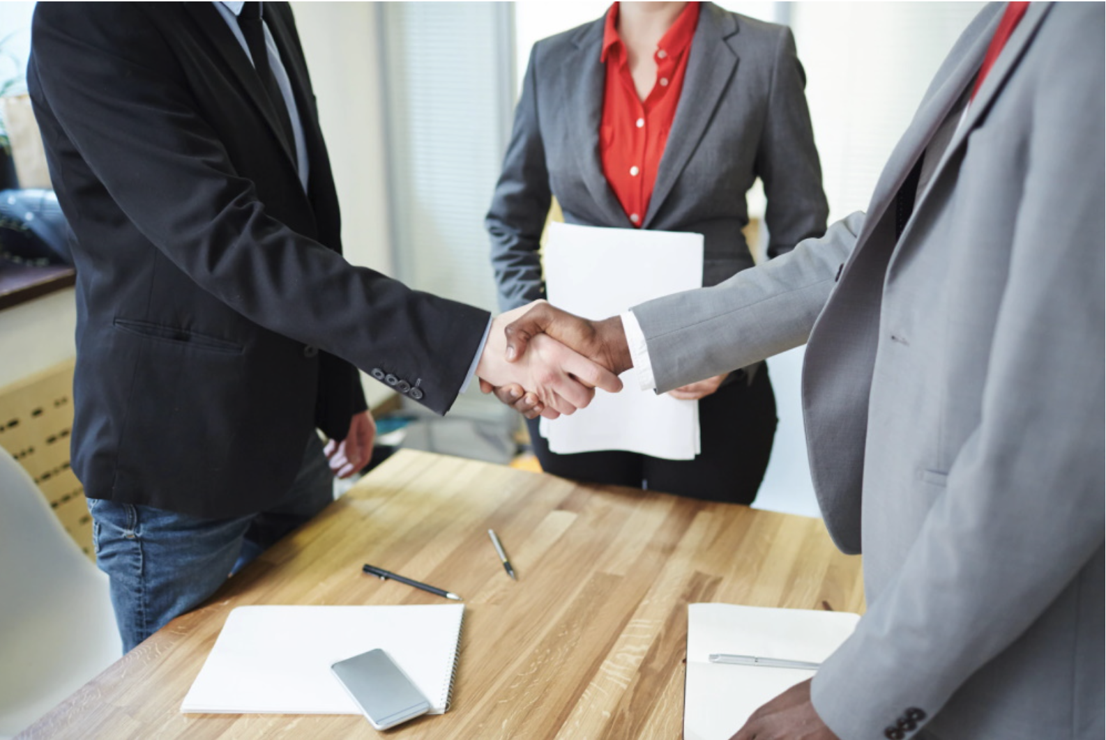Two people in business suits shaking hands over a wooden table, with a woman holding documents in the background.