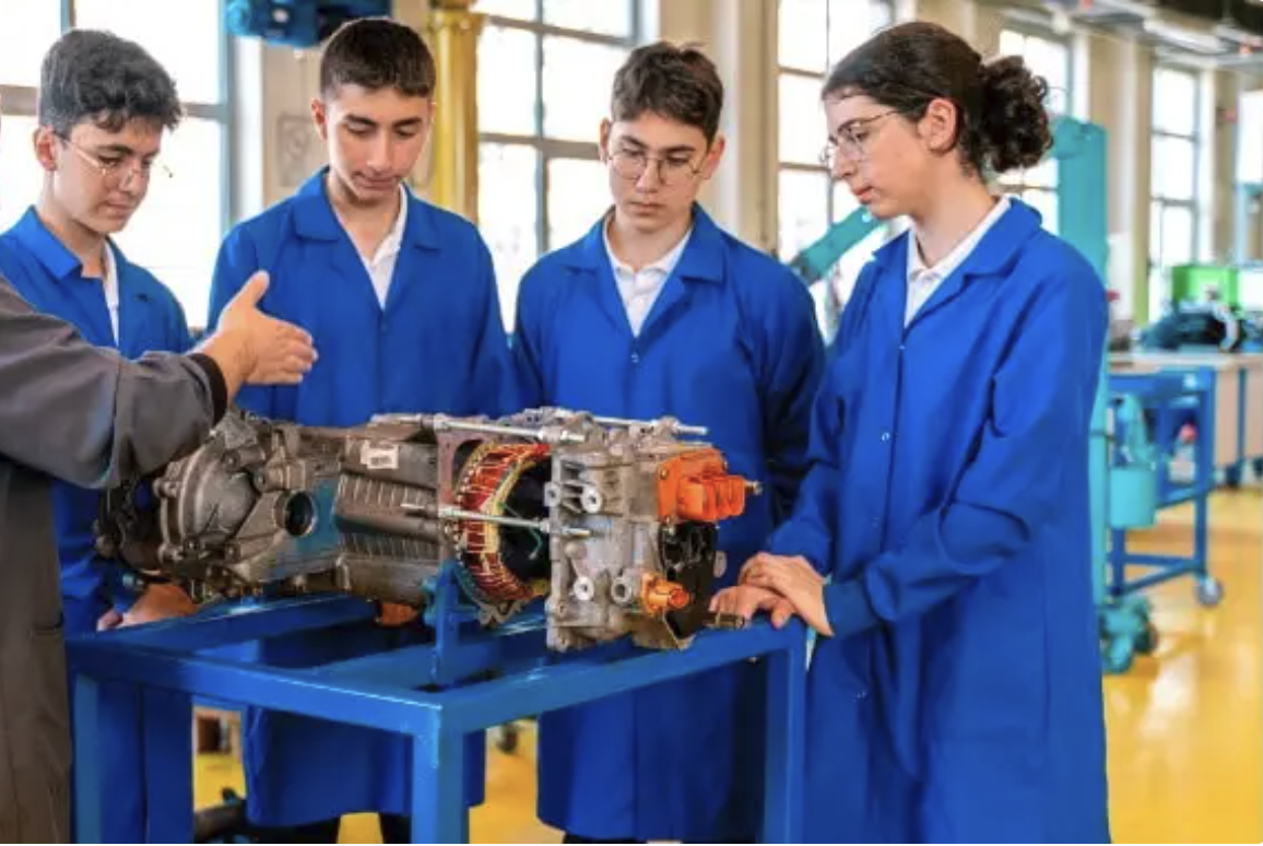 Four students in blue lab coats observing a mechanical device on a work table during a technical class or workshop.