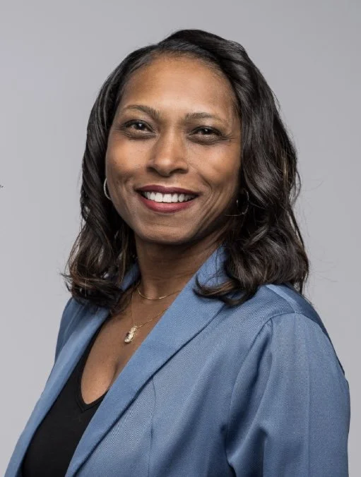 A smiling African American woman with dark shoulder-length hair, wearing a blue blazer over a black top, and a delicate necklace, against a gray background.