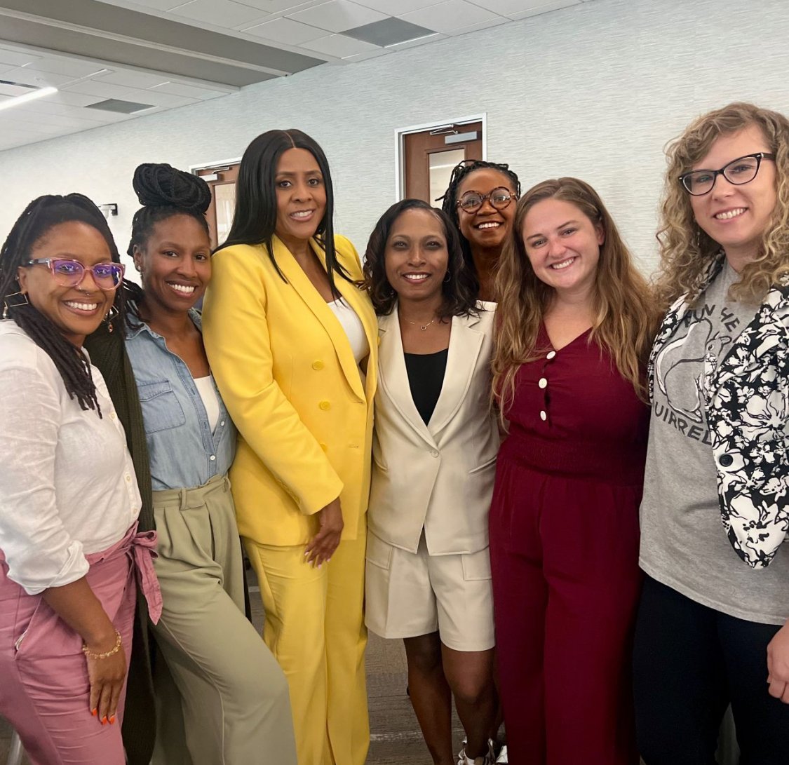 A modern wooden house situated on rolling green hills, with snow-capped mountains in the background under a cloudy sky.