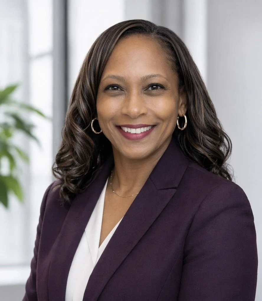A smiling African American woman with dark shoulder-length hair, wearing a blue blazer over a black top, and a delicate necklace, against a gray background.
