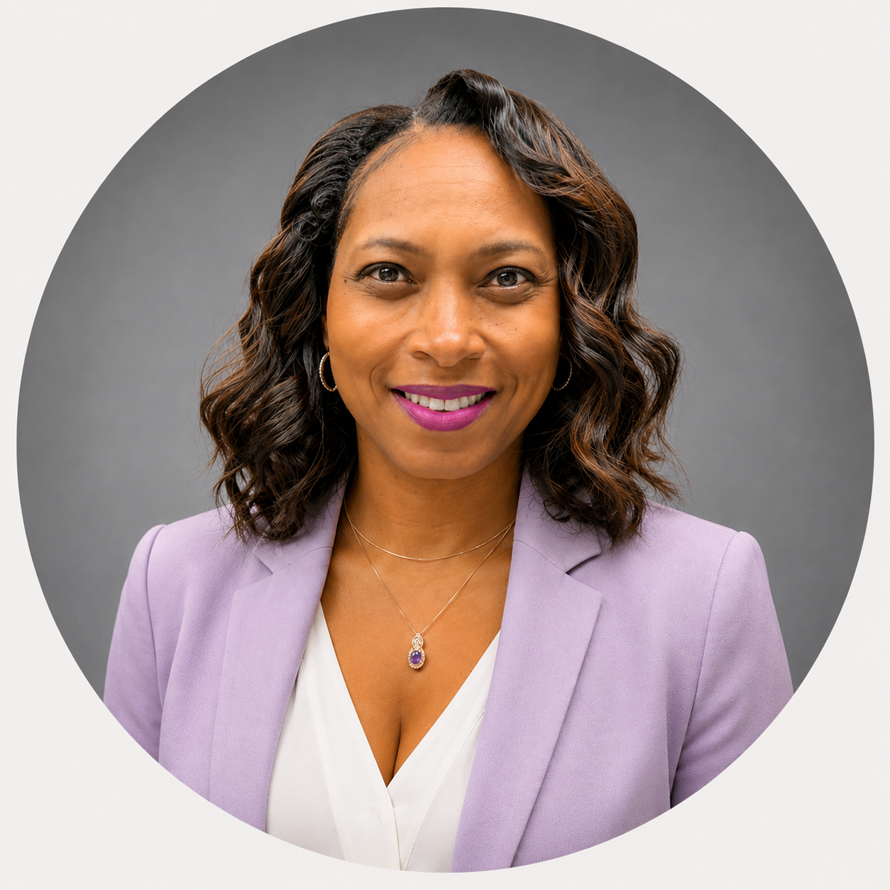 A smiling African American woman with dark shoulder-length hair, wearing a blue blazer over a black top, and a delicate necklace, against a gray background.