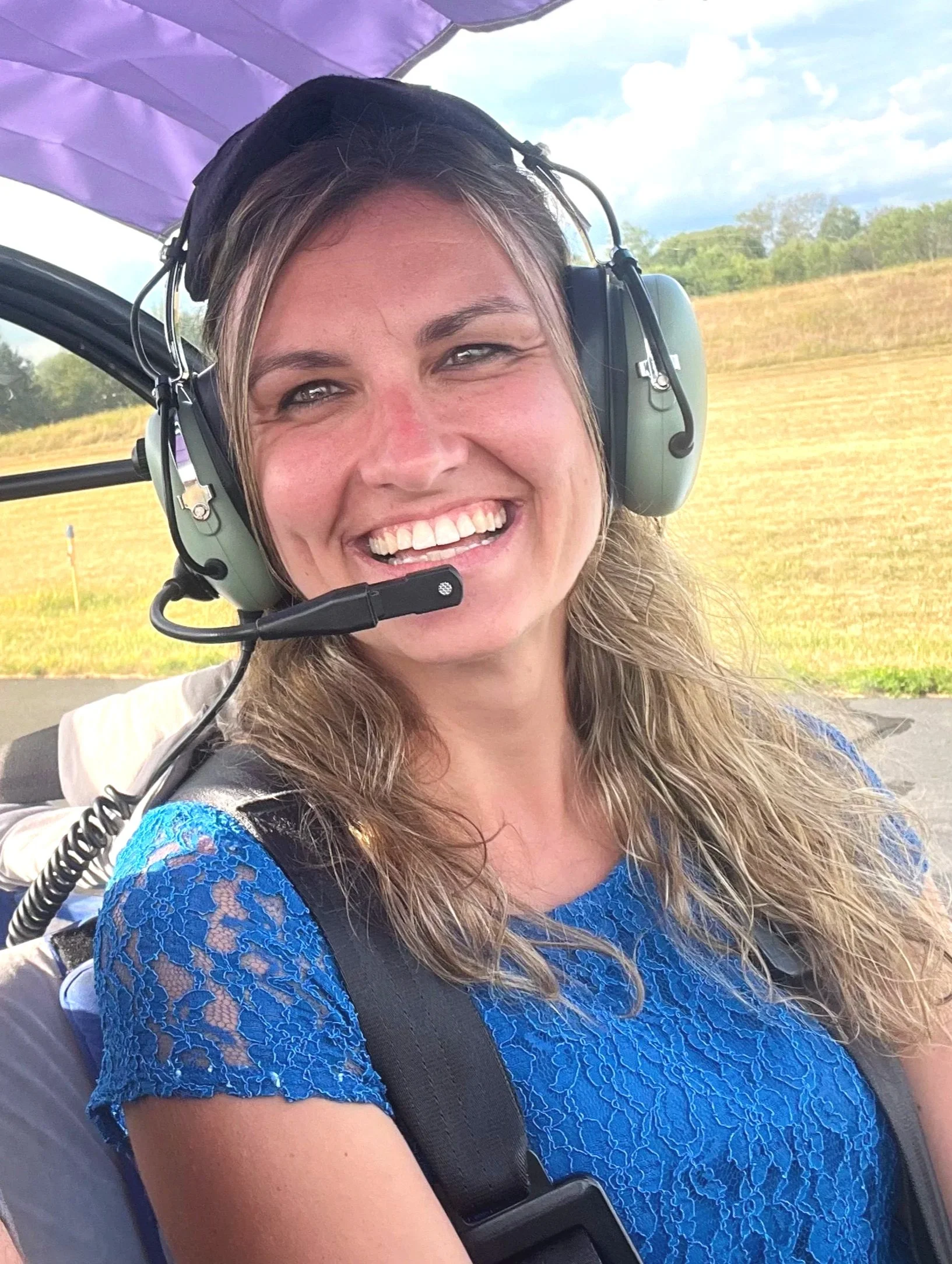 A woman with long wavy brown hair wearing a blue lace dress, smiling and wearing a headset with a microphone, standing outdoors under a purple canopy with a scenic grassy field and trees in the background.