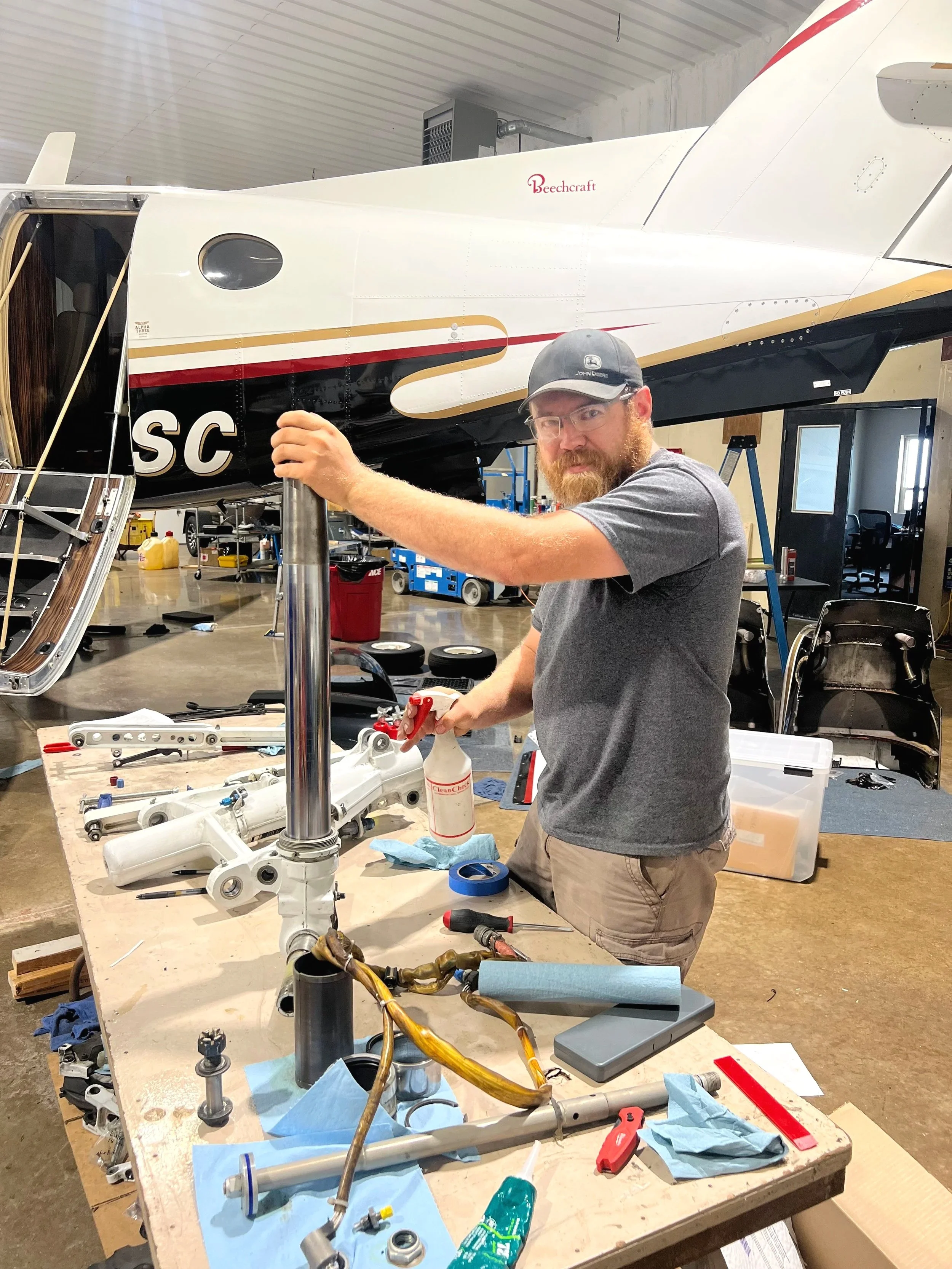 Man working on helicopter parts at a workbench inside a hangar, with a helicopter in the background.