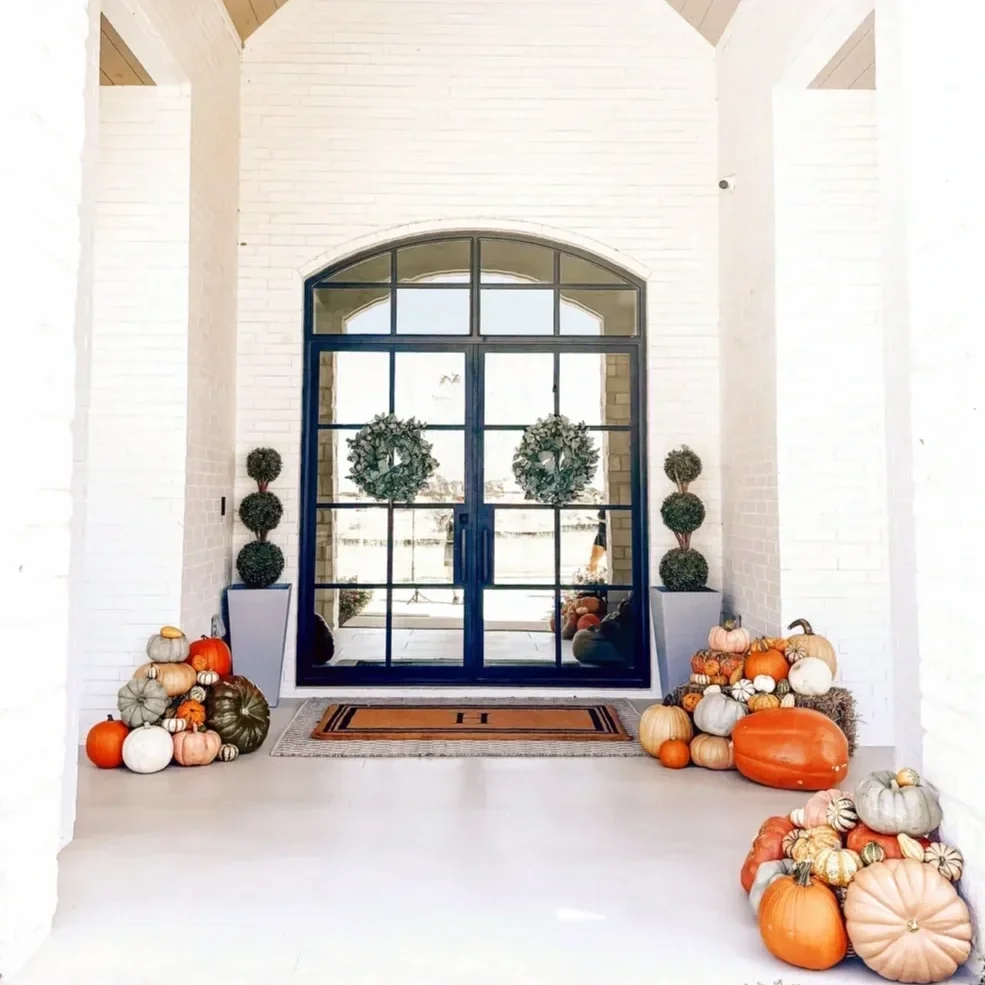 Decorative pumpkins and gourds arranged along the entryway floor and beside the door, with wreaths on the door, in a bright, white-walled entrance area.
