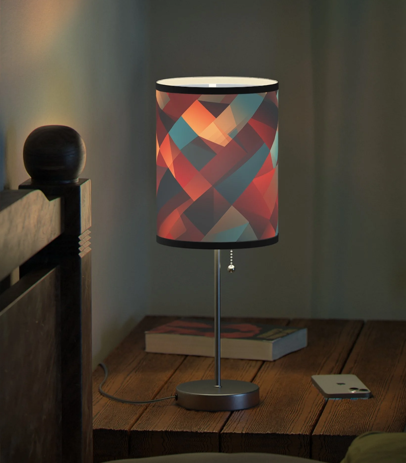 A decorative table lamp with a geometric patterned lampshade in warm colors, placed on a wooden surface next to a smartphone and a book.