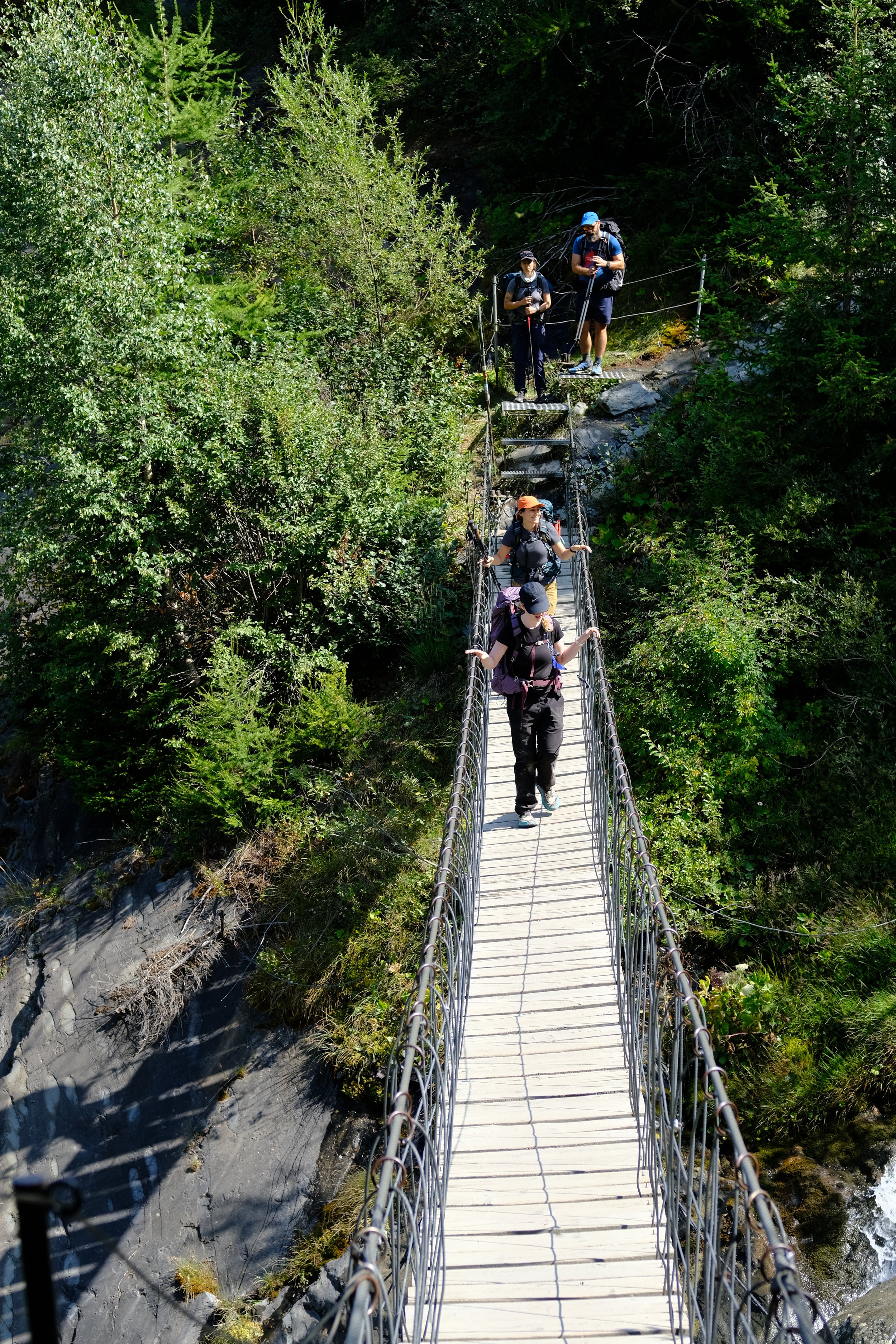Group of hikers walking across a narrow suspension bridge over a rocky stream in a wooded area.