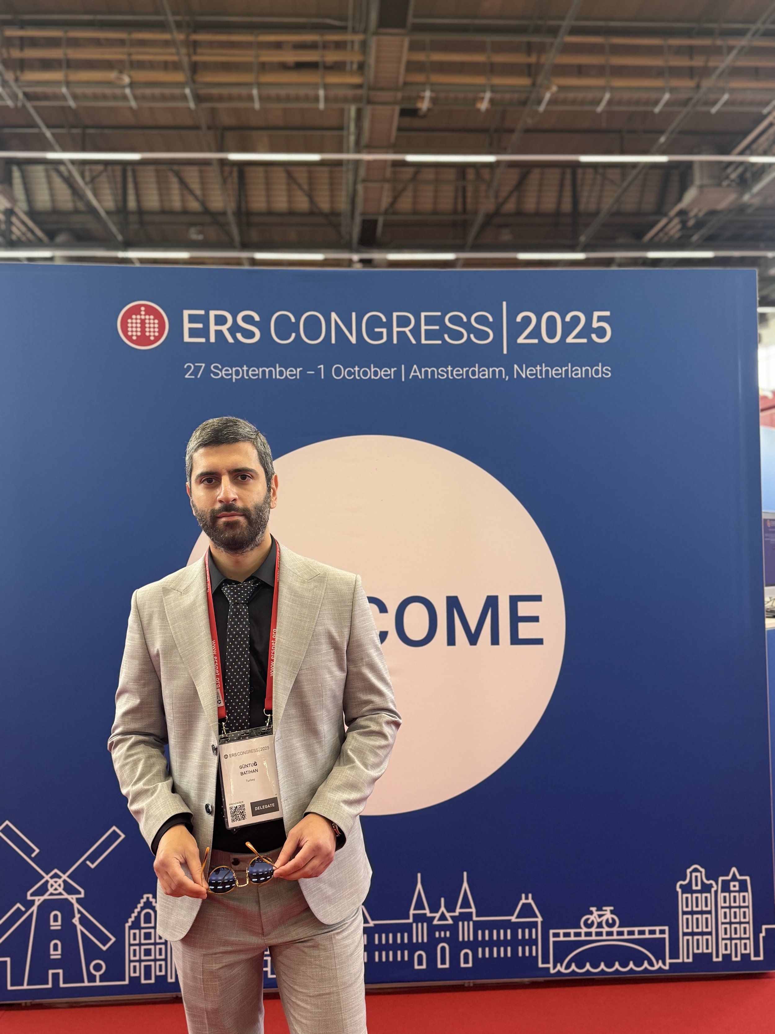 Amsterdam, Hollanda'da düzenlenen ERS Kongresi 2025 etkinliğine katılan erkek kişi. Kadınsız, gri takım elbise giymiş, kurumsal kimlik kartı taşıyor, arkasında büyük mavi bir kongre afişi var. Afişte kongre bilgileri ve şehir ile tarih bulunuyor.