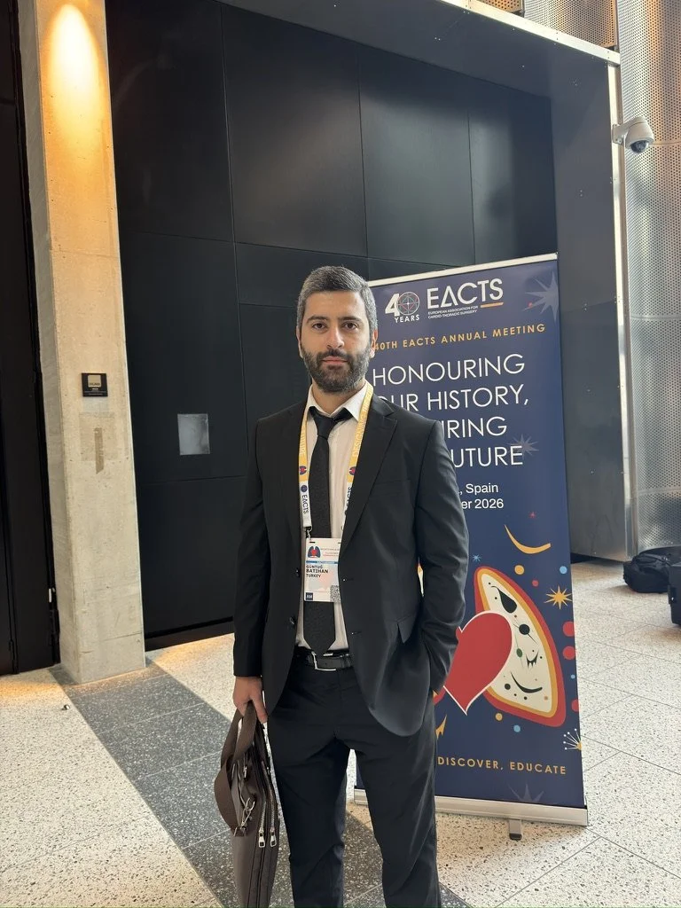 Erkek, siyah ceket ve kravat giymiş, elinde portföy tutuyor, ayakta ve arkasında parti temalı EACTS konferans banner'ı var.