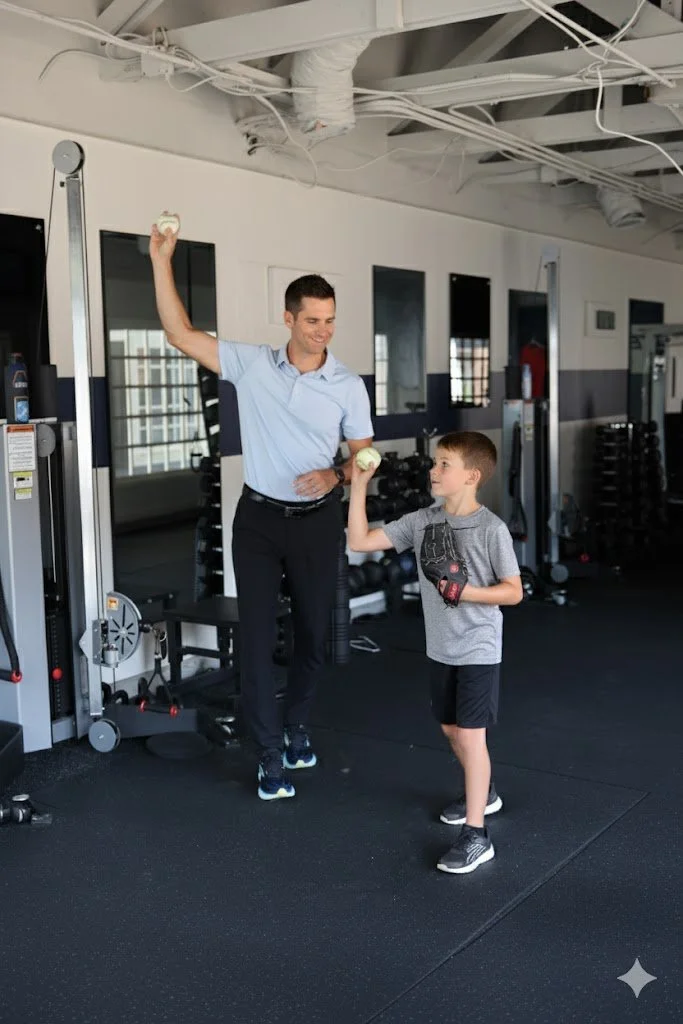 A baseball coach or trainer instructs a young boy in a gym, showing him how to throw a baseball. The boy is wearing a baseball glove, white shirt, blue shorts, and athletic shoes. The instructor is pointing towards something while demonstrating technique. The gym has weightlifting equipment and mirrors.