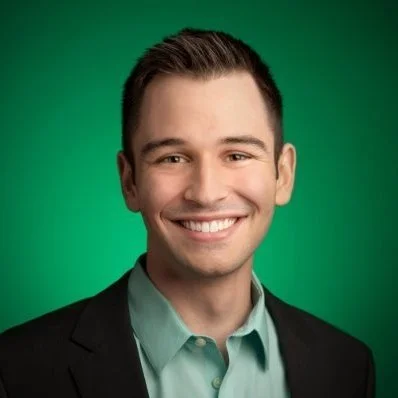 Headshot of a smiling man with short brown hair, wearing a black blazer and light green dress shirt, against a green background.