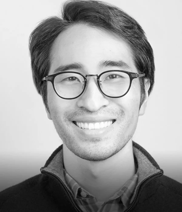 Black-and-white portrait of a smiling man with glasses and short hair, wearing a zip-up sweater over a collared shirt.