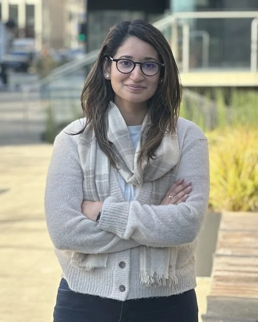 Young woman with glasses and long dark hair standing outdoors with arms crossed, wearing a beige cardigan, scarf, and white top, smiling slightly.