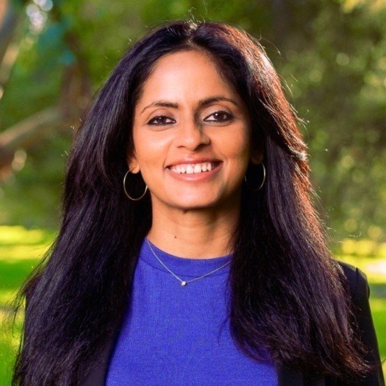 Smiling woman with long dark hair, wearing hoop earrings, a blue top, and a necklace outdoors with green trees in the background.