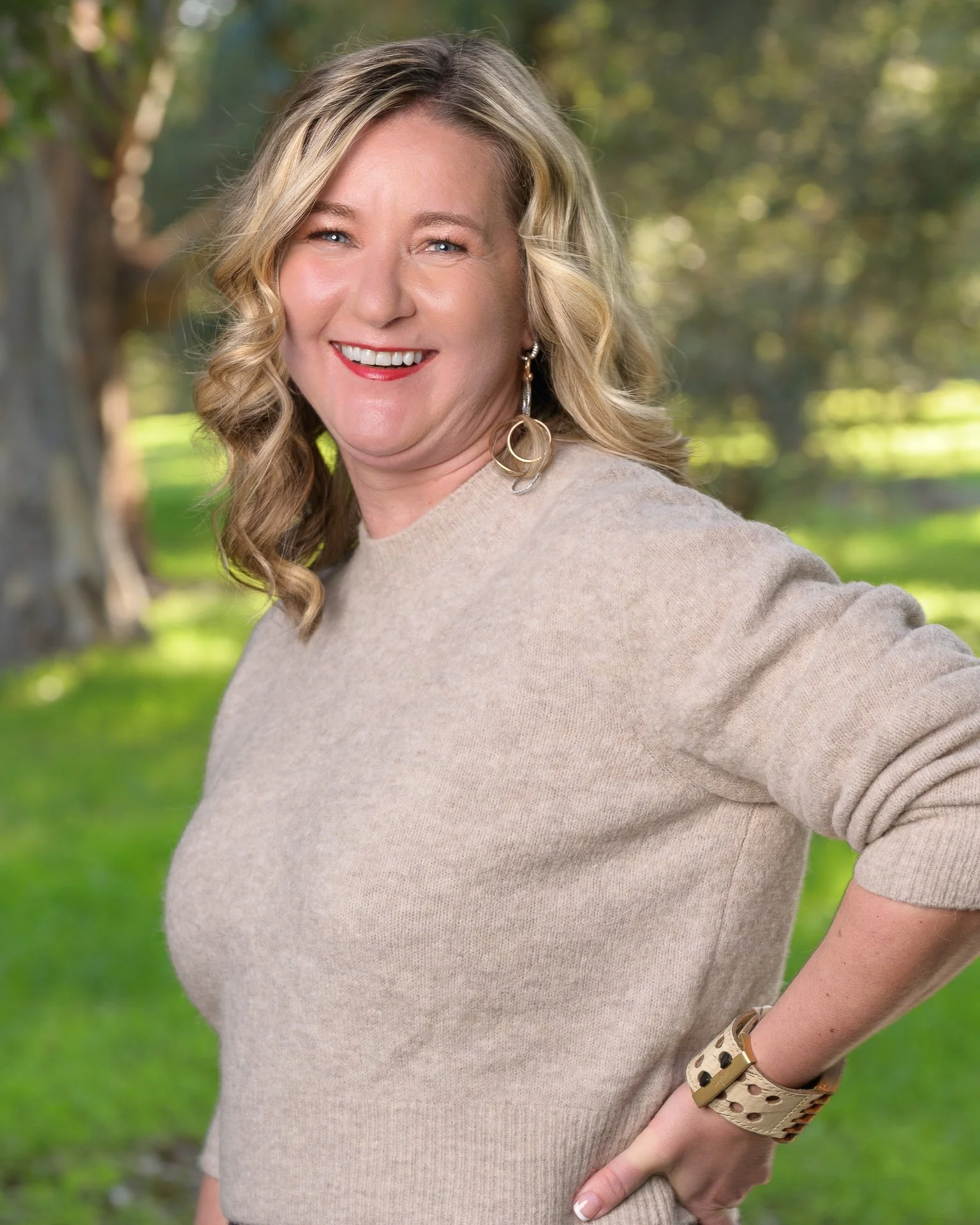 A woman with blonde hair styled in loose curls, smiling outdoors with a background of trees and greenery. She is wearing a beige sweater, large hoop earrings, and a wide bracelet, with one hand on her hip.