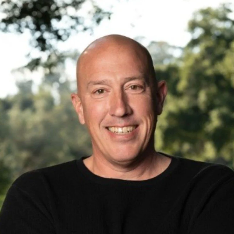 A smiling man with a shaved head wearing a black shirt outdoors, with trees and greenery in the background.