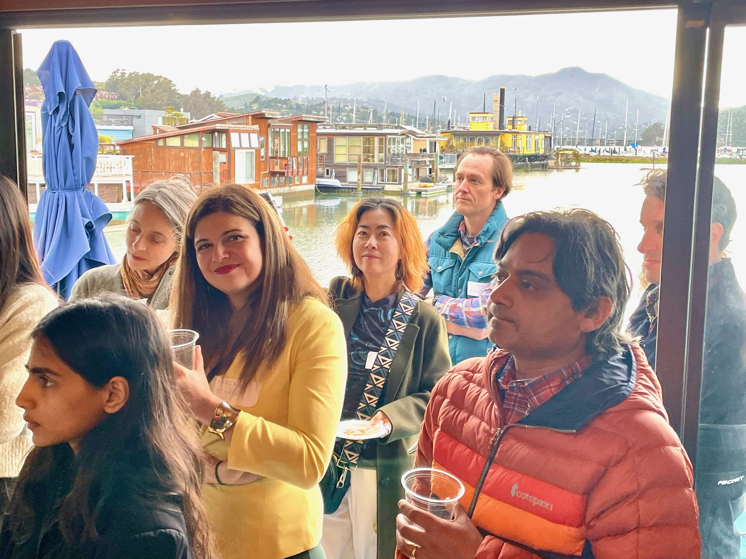 Group of diverse people on a boat patio, with a view of boats and houseboats in the harbor and mountains in the background.
