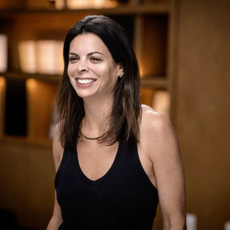 A woman with shoulder-length dark hair, smiling, wearing a black sleeveless top, standing in a warmly lit room with a bookshelf in the background.