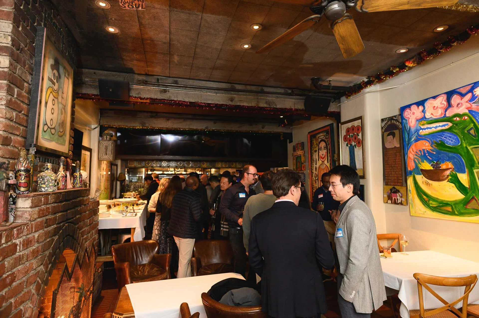 People gathered in a restaurant with colorful walls decorated with paintings and artwork, including a large vivid painting of a crocodile on the right.