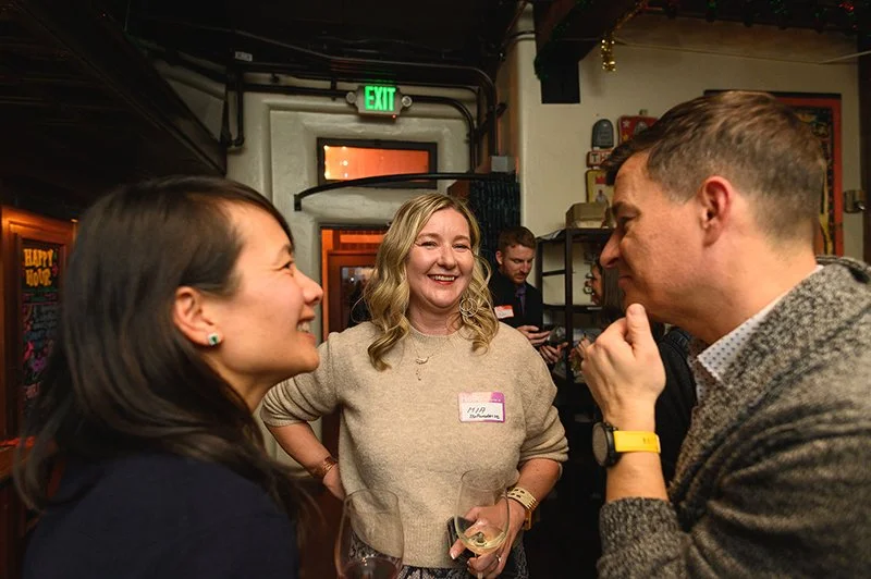 Three people smiling and talking at a social gathering in an indoor bar or restaurant, with one holding a glass of white wine.