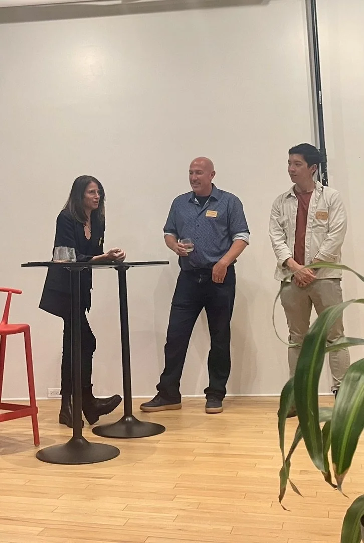 Three people standing together near a tall table, engaged in conversation, in a room with a plain white wall and wooden floor, one holding a glass of wine.