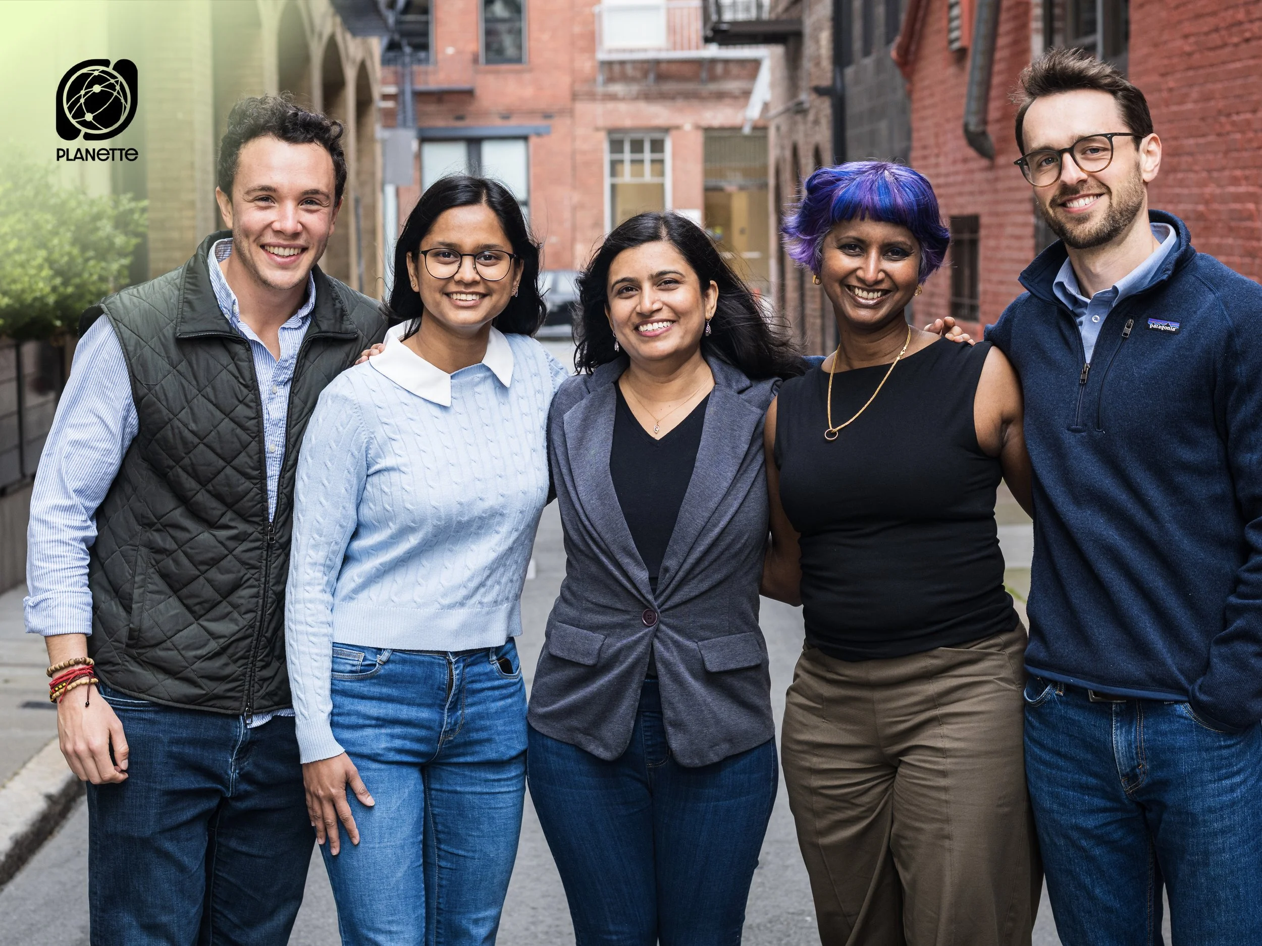 A diverse group of five people standing closely together outdoors, smiling, with arms around each other, in an alleyway with red brick buildings.