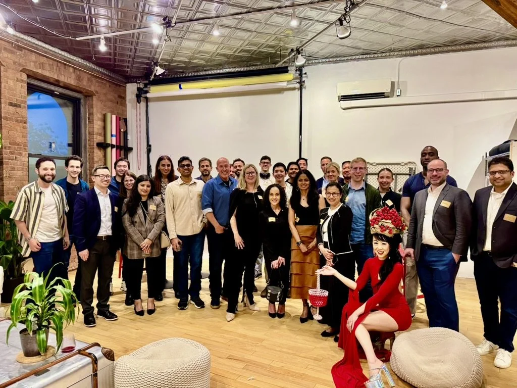 Group of people gathered in an indoor space, posing for a photo, with a woman dressed in a red outfit and headpiece sitting in front.