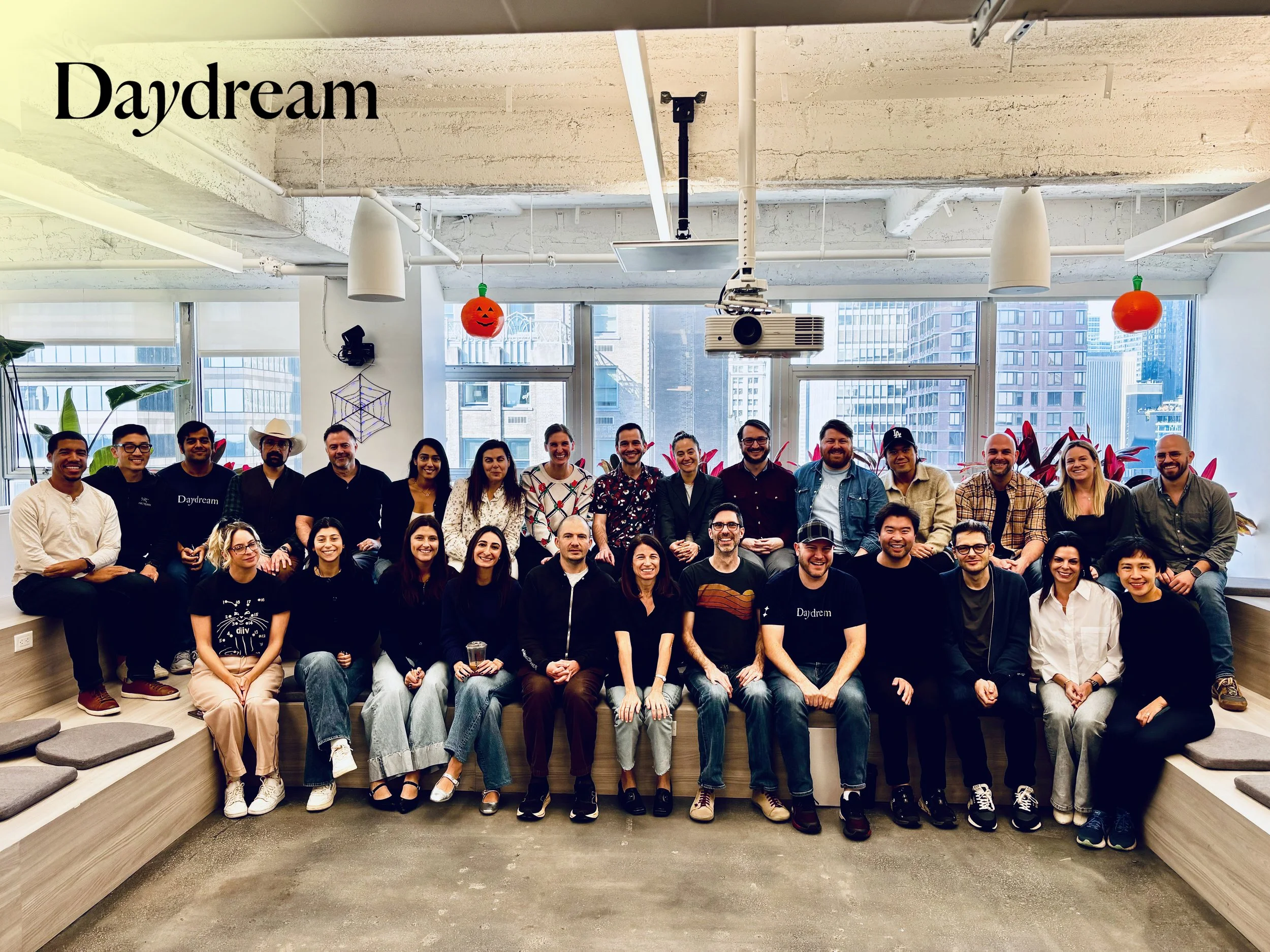 A group of people gathered in an office space, sitting and standing on a wooden platform, with windows showing city buildings in the background. The room is decorated with floating pumpkin and apple decorations and a spider web. The word 'Daydream' is written in black text in the upper left corner.