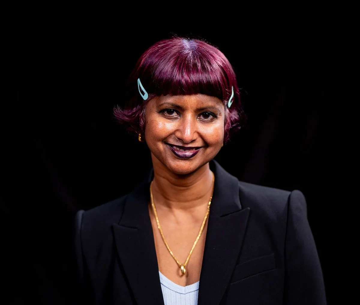 A woman with short burgundy hair styled with hair clips, wearing a black blazer and gold jewelry, smiling against a black background.