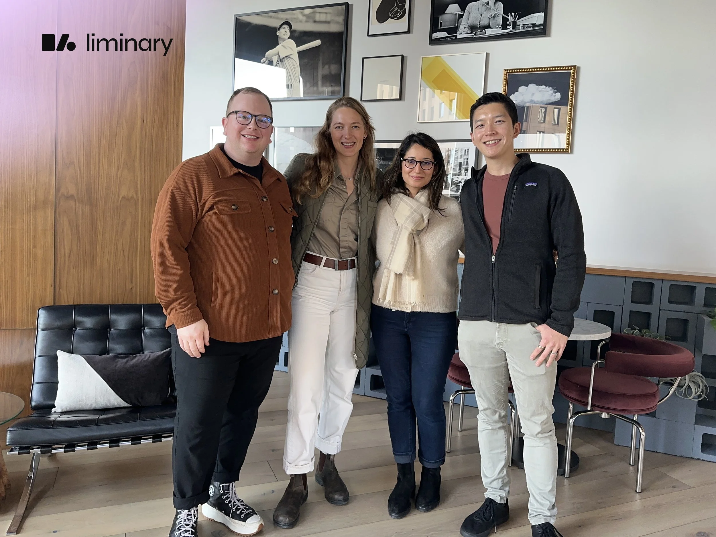Four people standing inside a modern room, smiling and posing for a photo. The room features framed artwork on the wall, a wooden panel, a black chair with a patterned pillow, and a small round table with red chairs.