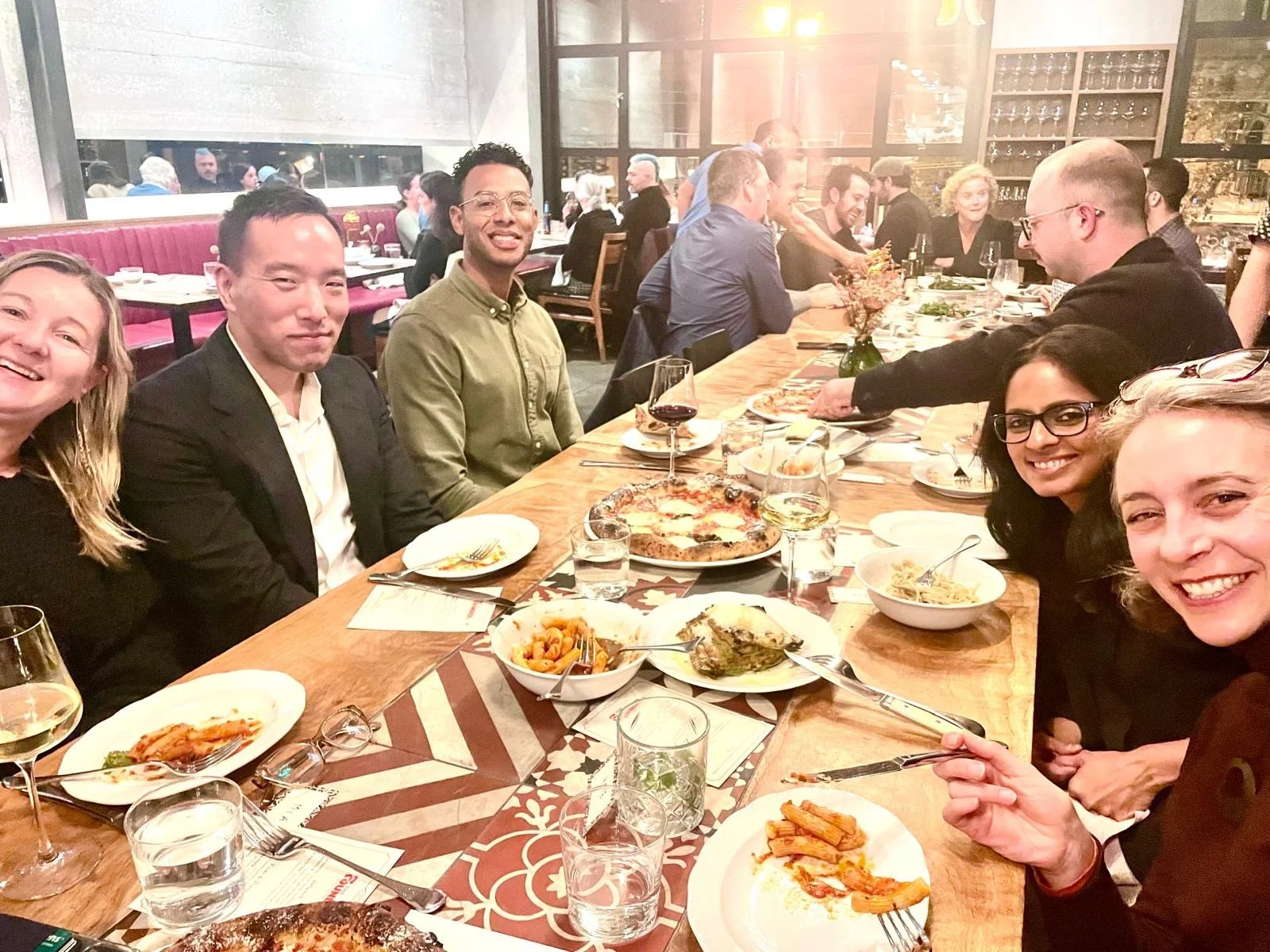 Group of friends enjoying dinner at a restaurant, sitting around a wooden table with pizza, pasta, and drinks.