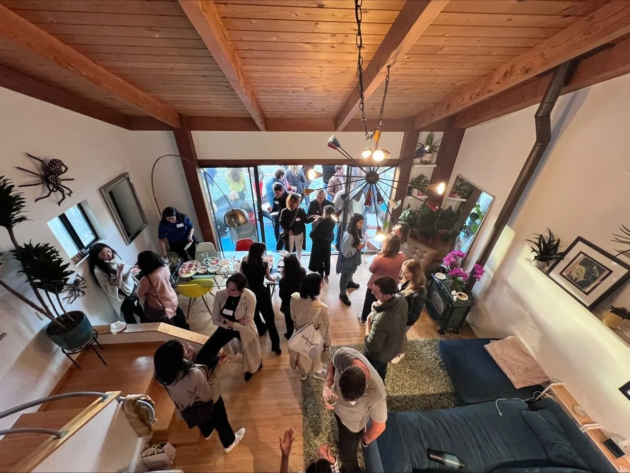 People attending a gathering inside a home with wooden ceiling, potted plants, and a chandelier, some near the window and others socializing or standing around.