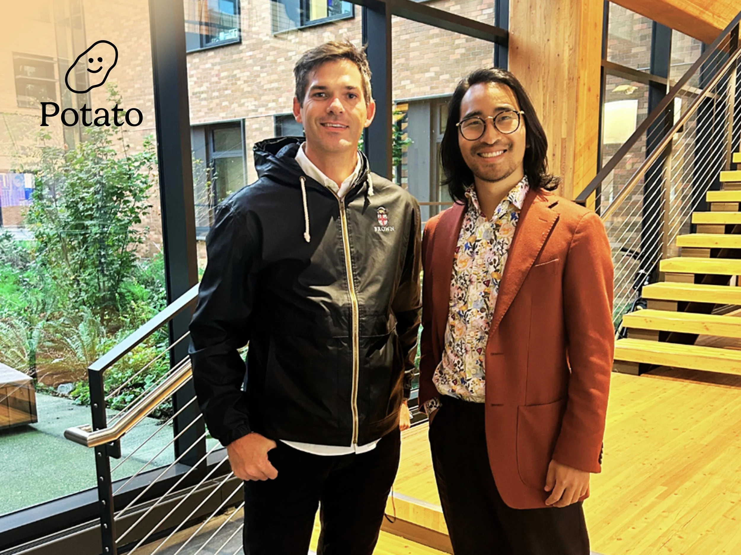 Two men standing inside a building with large windows, displaying a garden outside. One man is wearing a black jacket with a hoodie, and the other is wearing a burnt orange blazer with a patterned shirt. There is a hand-drawn potato graphic with the word "Potato" next to it in the upper left corner of the image.