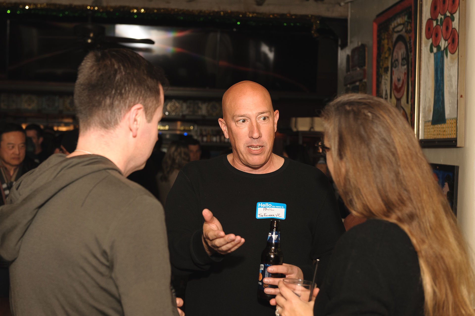 Three people engaged in conversation at a social event, with a man in the center wearing a name tag, holding a beer, and talking to a man and woman on either side of him, in a dimly lit room decorated with colorful artwork.