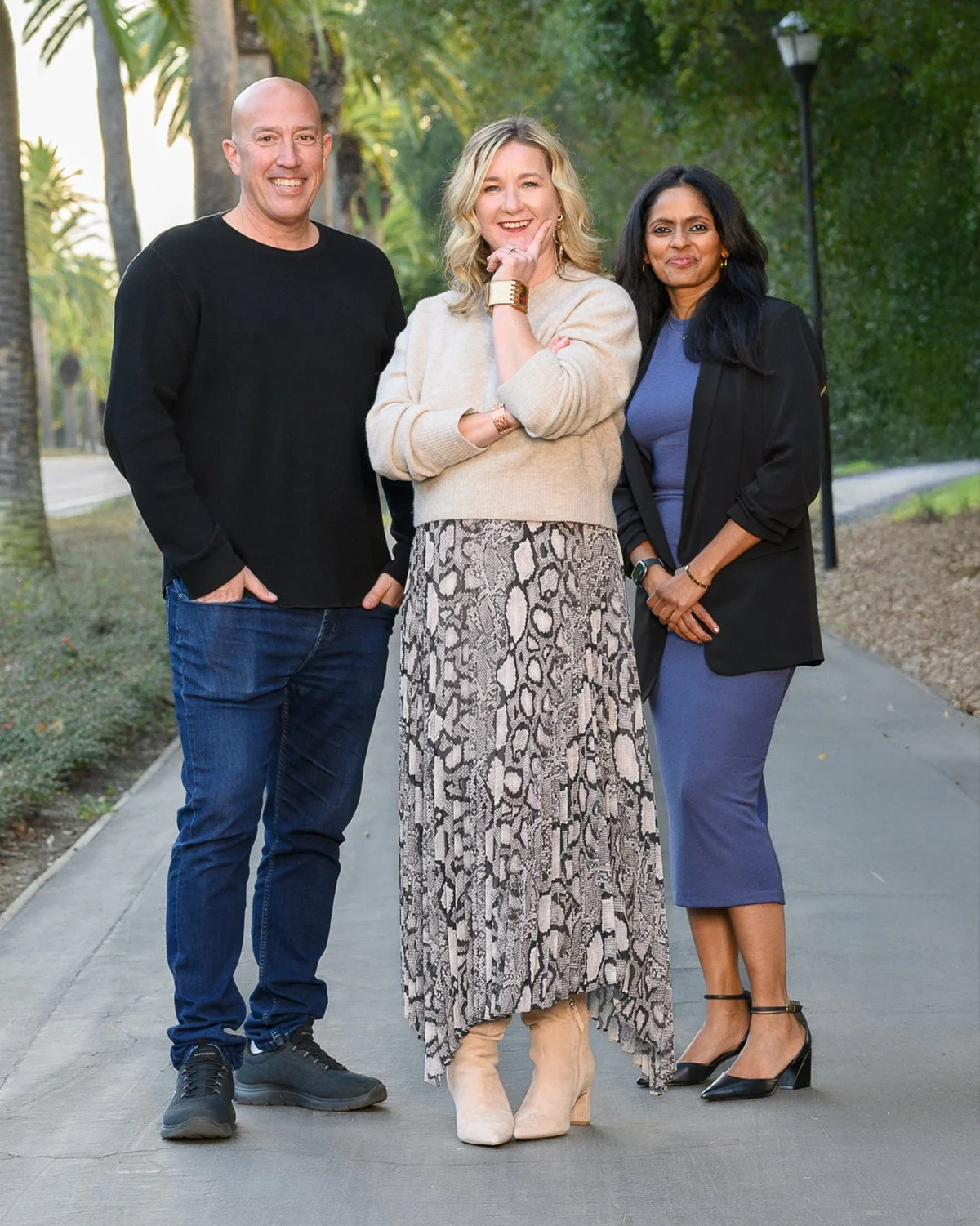 Three people standing outdoors on a sidewalk with trees and street lamps in the background, smiling at the camera.