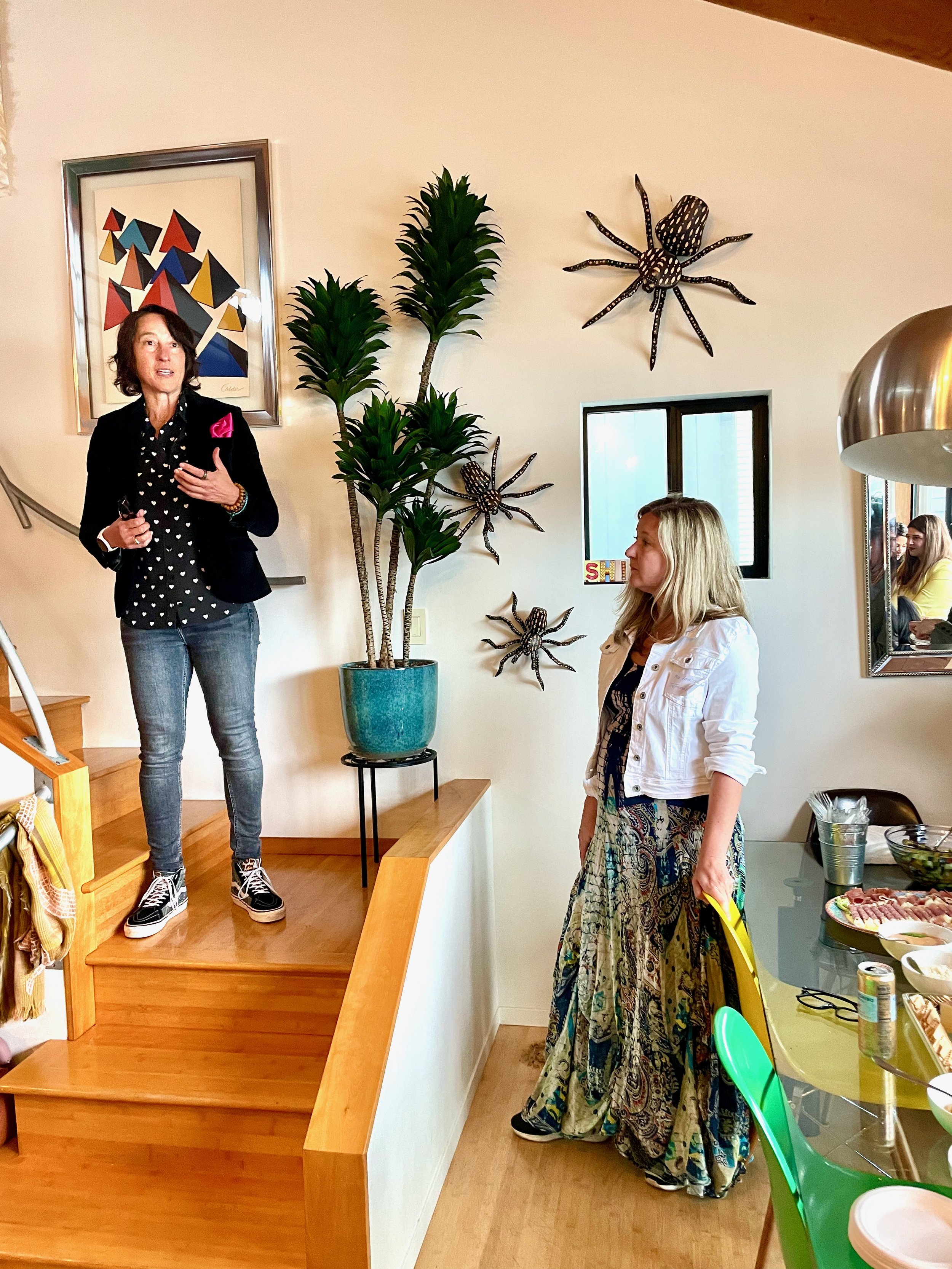 A woman standing on a small wooden staircase speaking to another woman standing below near a table with food. The woman on the stairs has short dark hair, is wearing a black blazer, gray jeans, and sneakers. The woman below has long blonde hair, a white jacket, and a colorful patterned skirt. There is a large potted plant, framed artwork, and decorative spider wall art on the wall behind them.