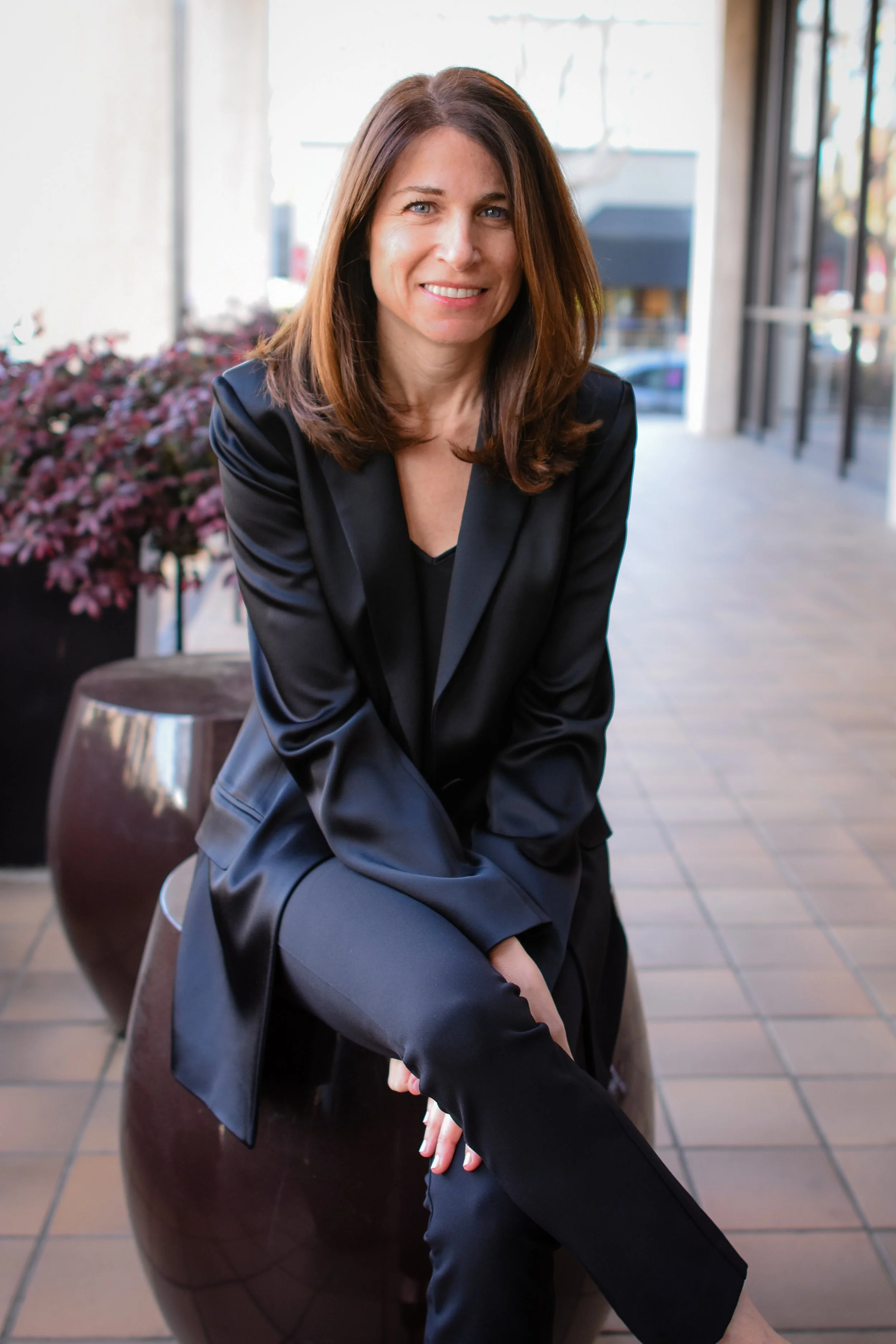 A woman with shoulder-length brown hair wearing a black suit, sitting on a brown ceramic stool outside on a sidewalk with plants and storefronts in the background.