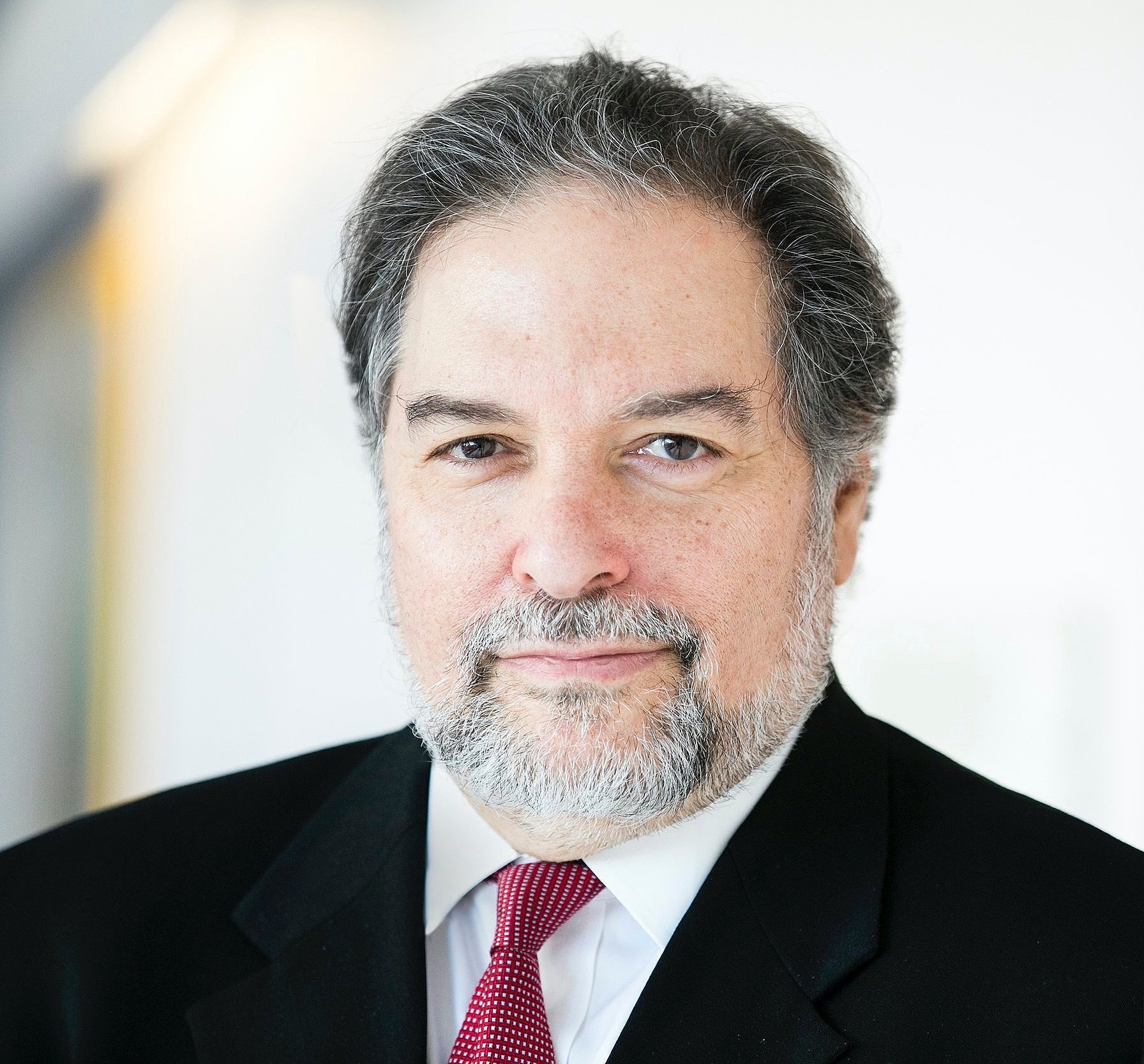 Close-up of a middle-aged man with graying hair and beard, wearing a black suit, white shirt, and red tie, looking at the camera with a slight smile.