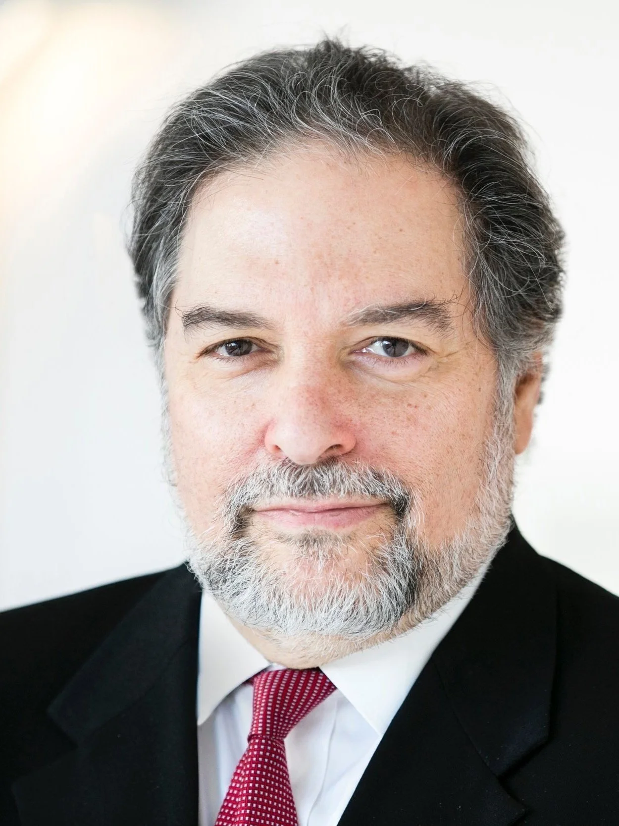 Close-up portrait of a middle-aged man with salt-and-pepper hair and beard, wearing a black suit, white shirt, and red tie, looking at the camera with a slight smile.