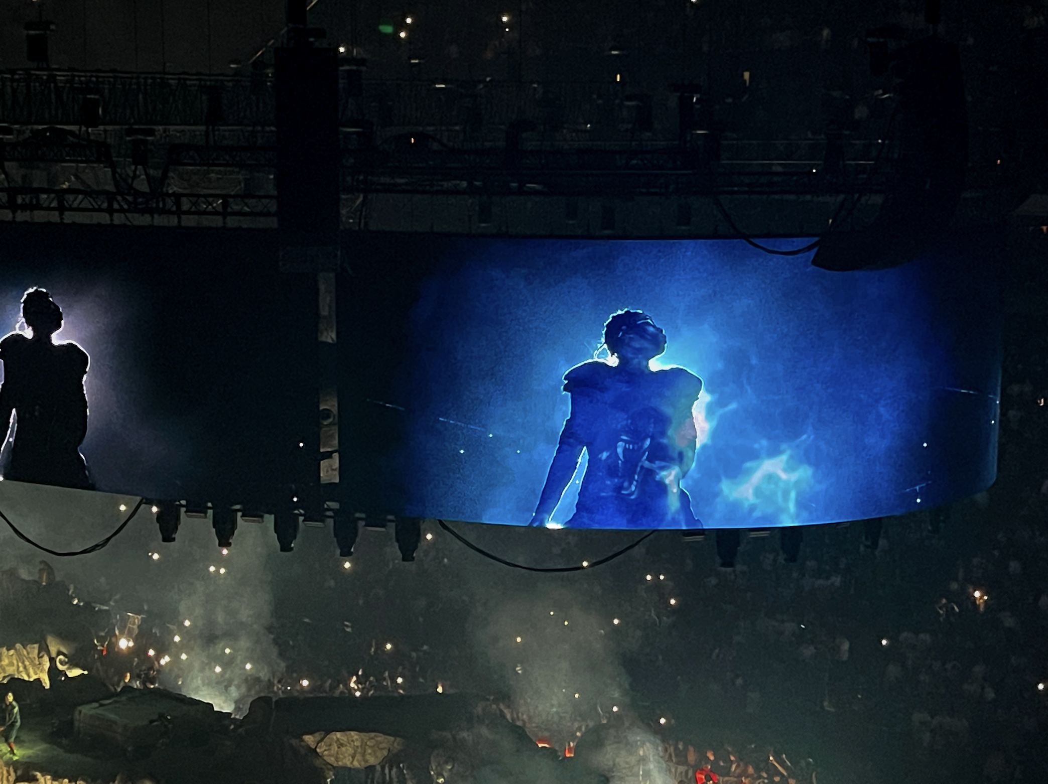 Large digital screen at concert showing Travis Scott with a glowing blue aura, surrounded by dark silhouettes of audience members and stage equipment.
