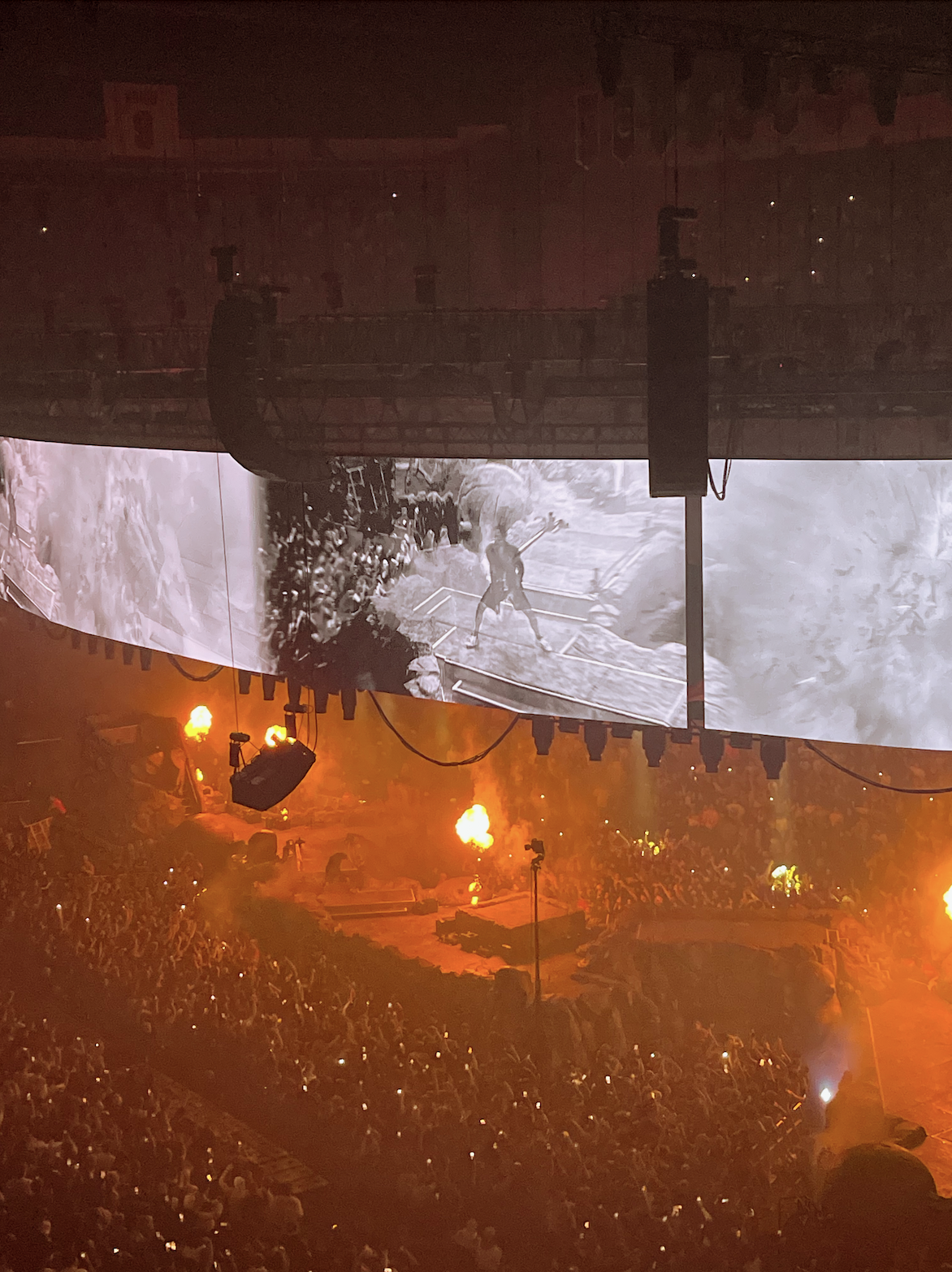 A large concert arena filled with a cheering crowd, with flames and fireworks on stage, and a giant high-definition screen displaying a black and white image of Travis Scott.