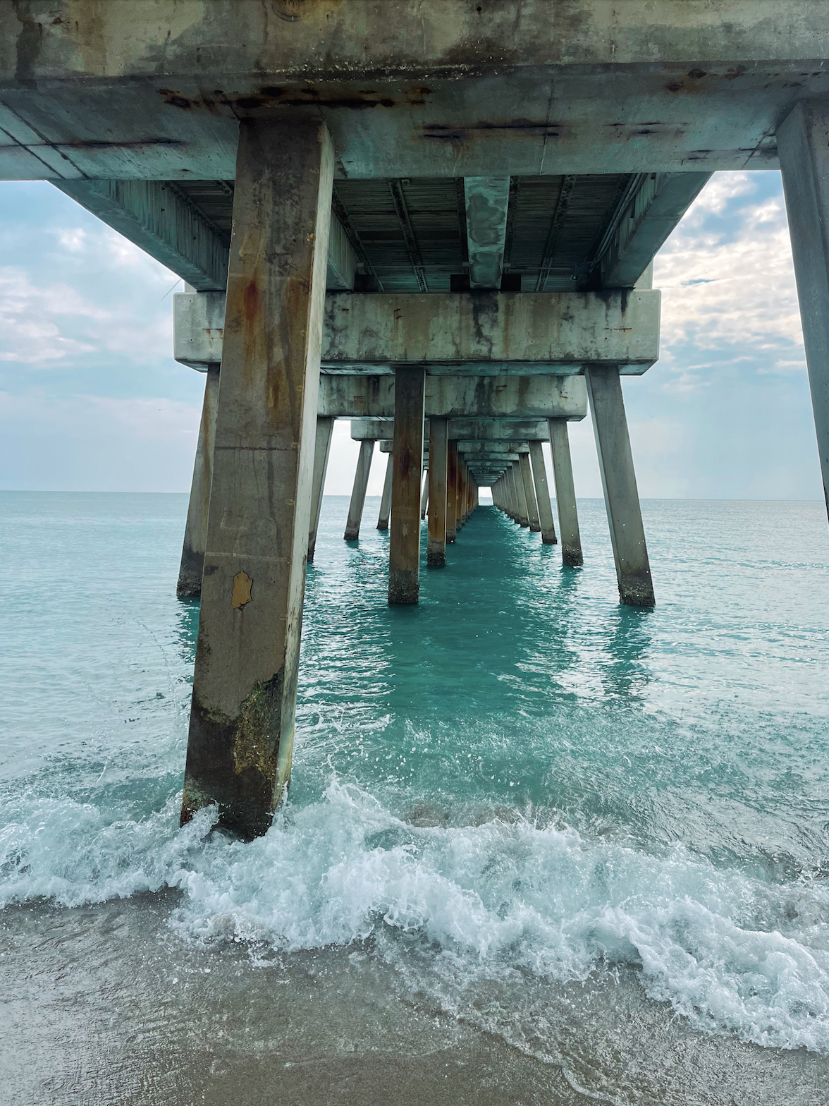 Underneath view of a weathered pier extending into the ocean with turquoise water and partly cloudy sky.