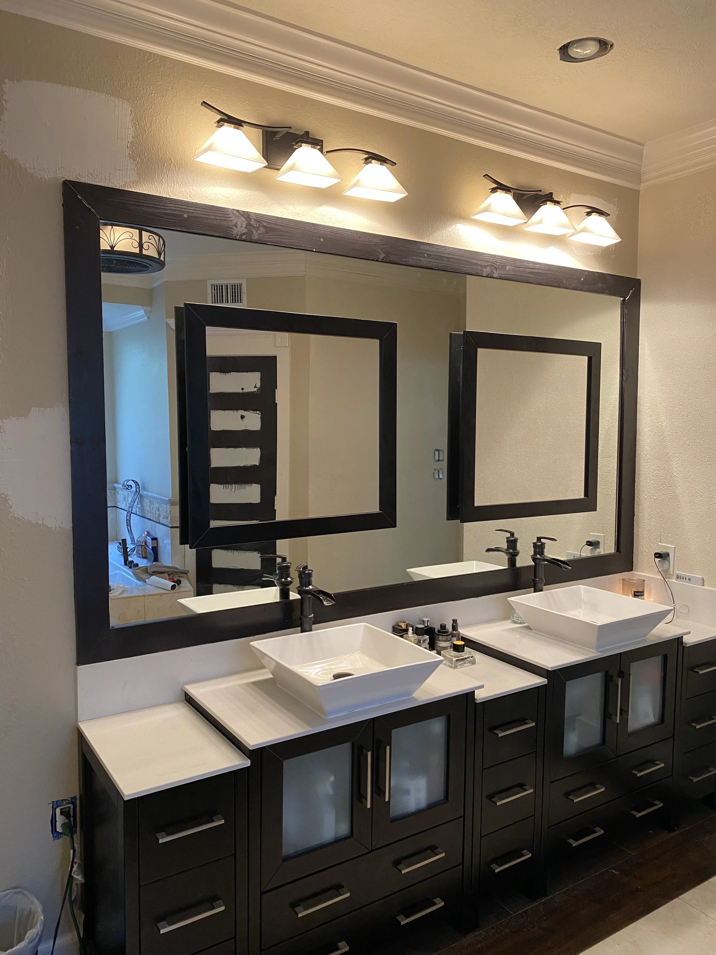 Double bathroom vanity with two white vessel sinks, black cabinets, large framed mirror, and three-light overhead fixture in a bathroom under renovation.