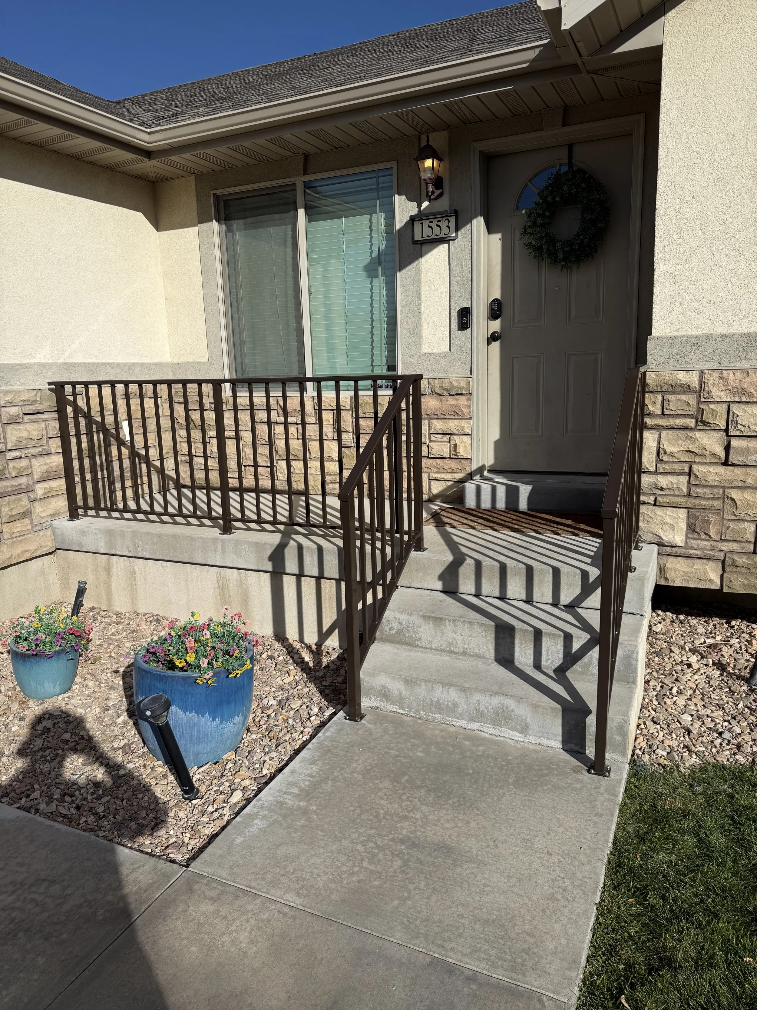 Front entrance of a house with a small concrete porch, metal railing, potted flowers, and a door with a wreath. The house number 1553 is displayed beside a wall-mounted lantern.