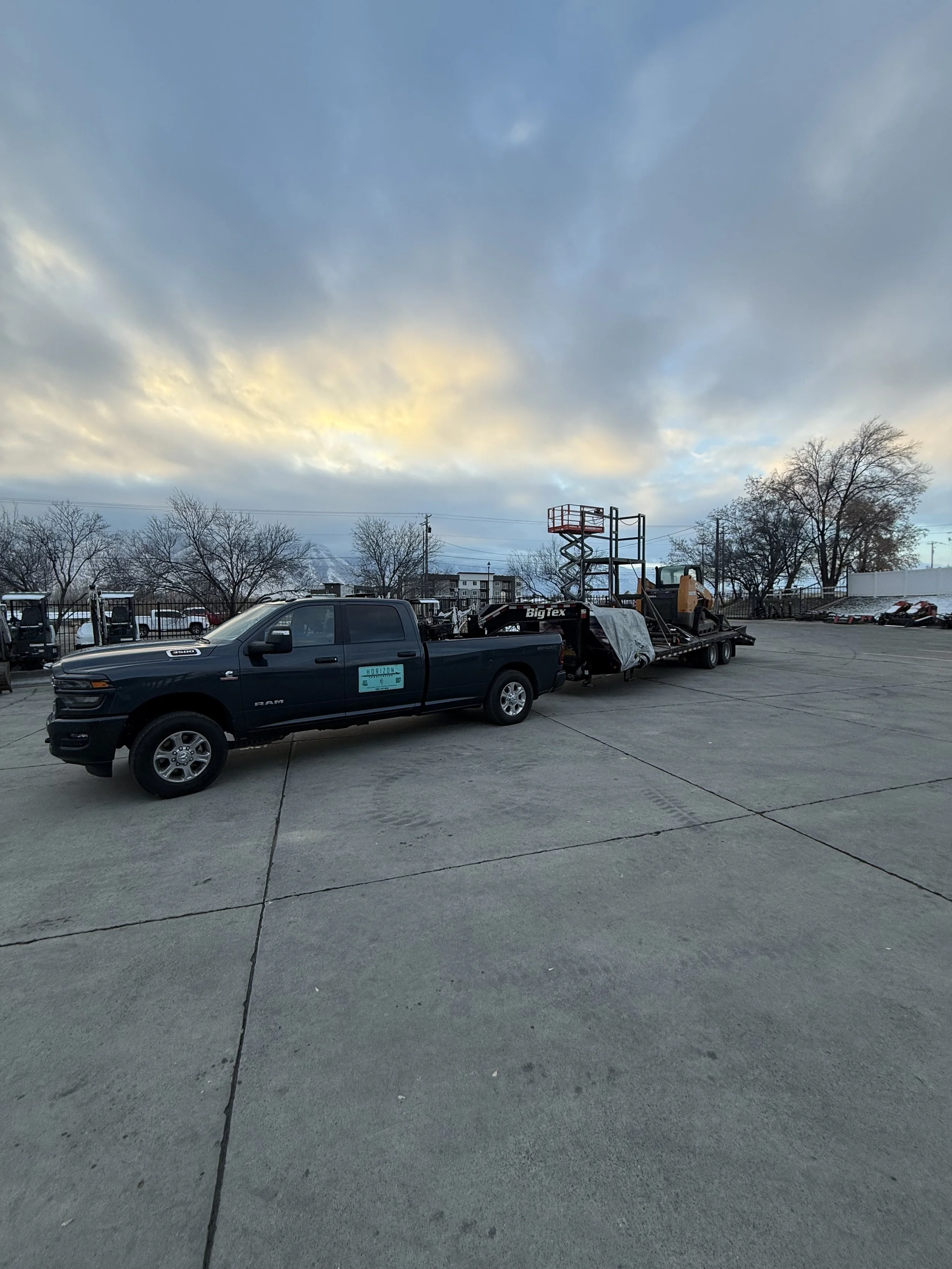 A black pickup truck parked in a large empty parking lot with equipment and a construction lift trailer attached, under a partly cloudy sky at dusk.