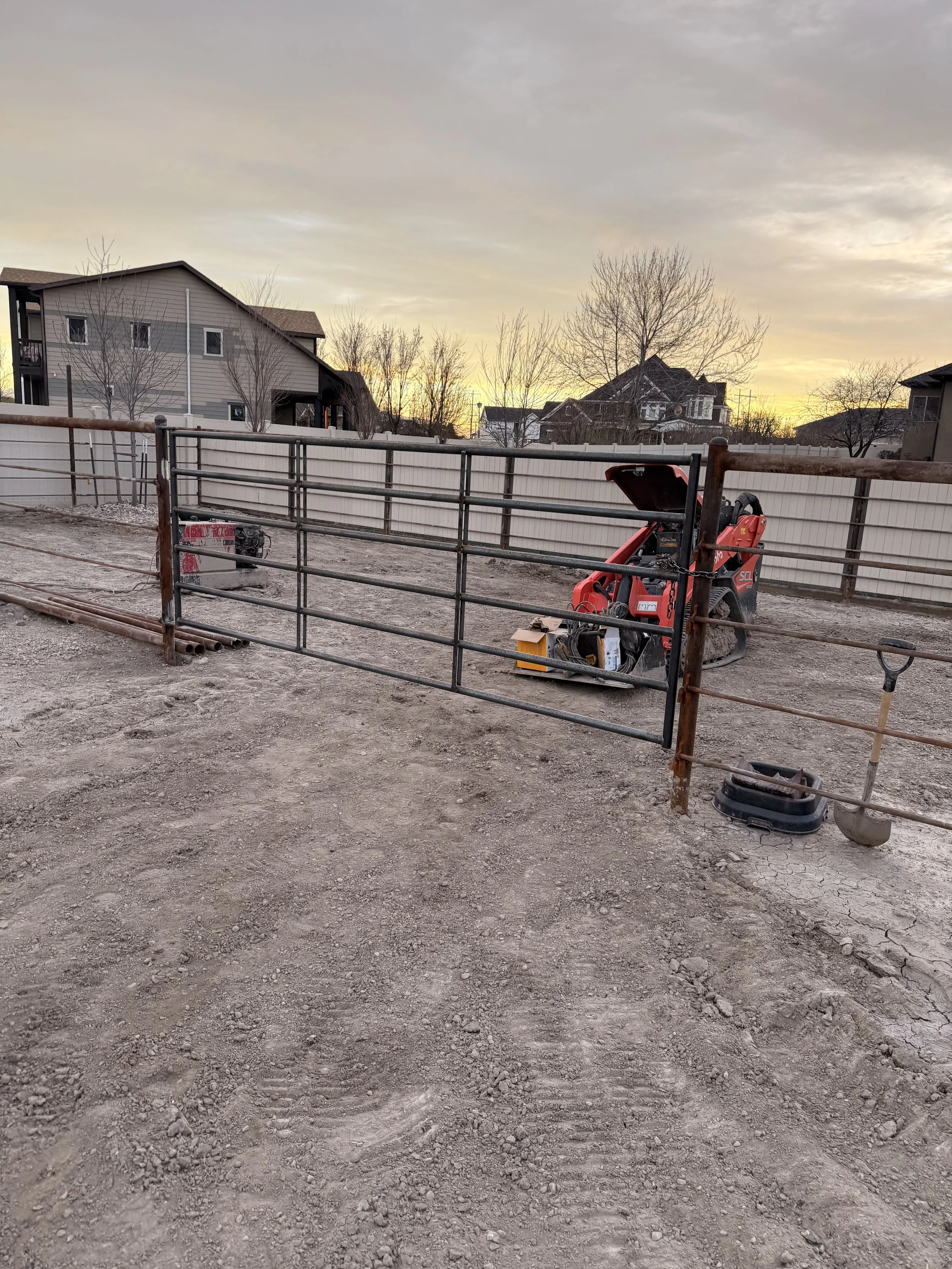 A fenced backyard with construction equipment, including a small red skid-steer loader, a shovel, and a bucket, on dirt ground during sunset.
