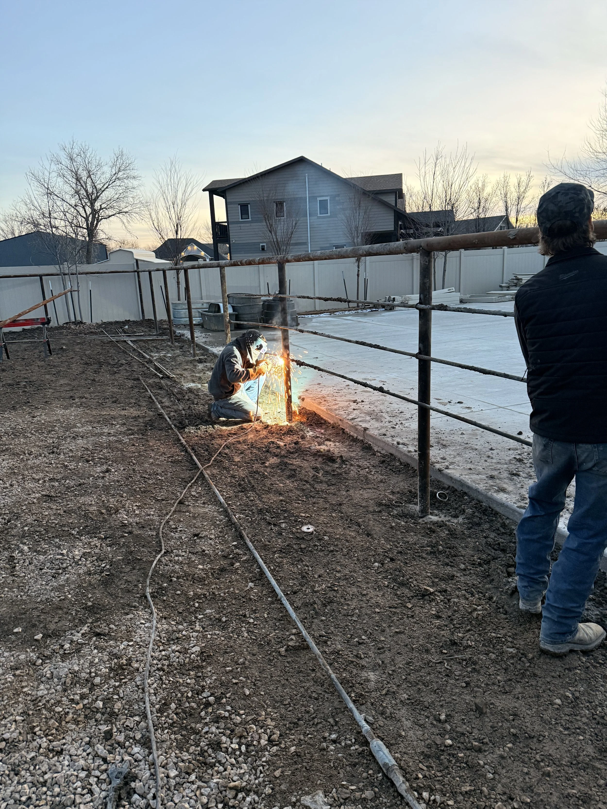Two workers welding metal pipes in a backyard, with one kneeling and welding, the other standing nearby, during sunset.