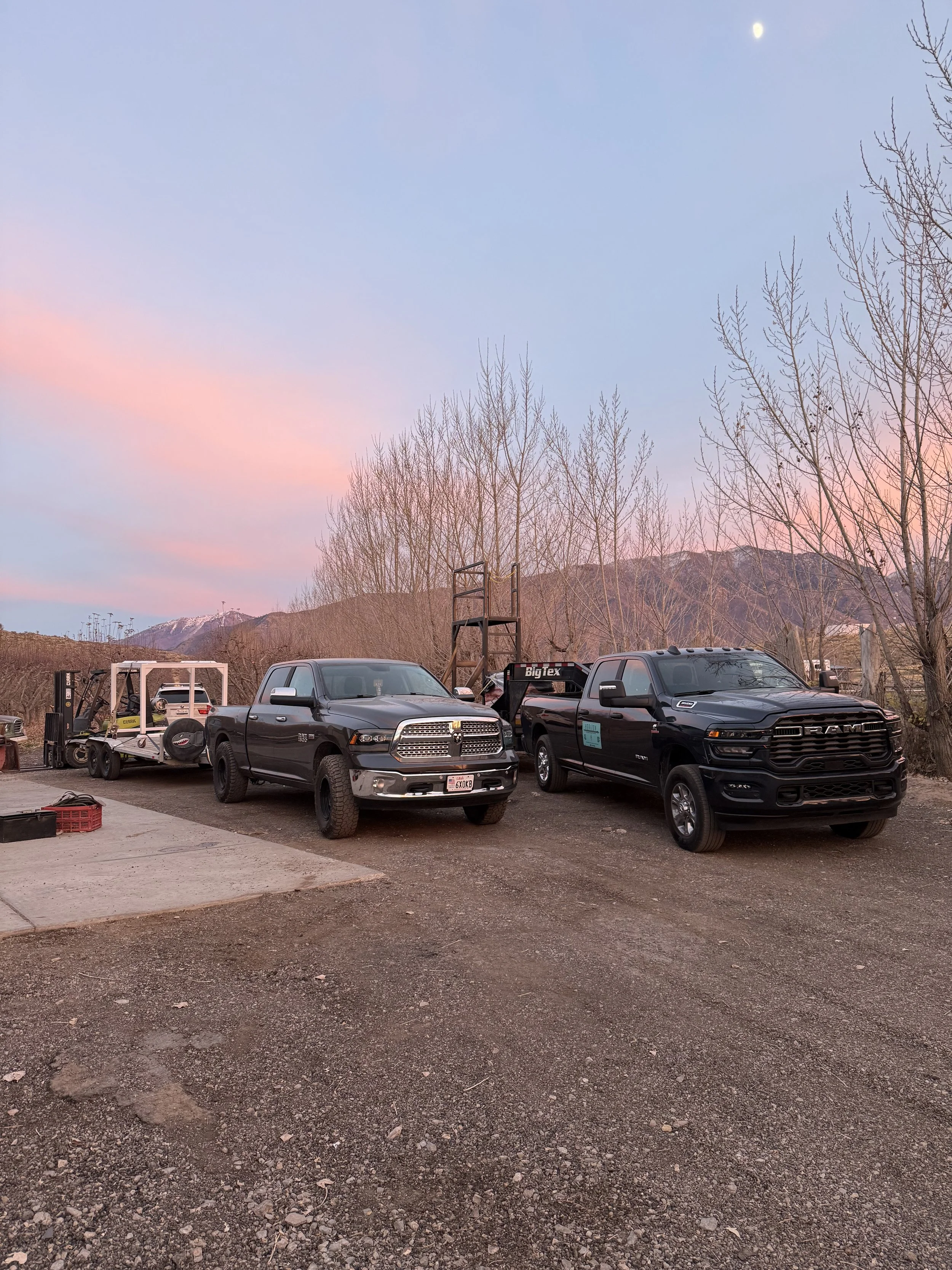 Two black pickup trucks, one hauling a trailer with equipment, parked on dirt ground during sunset. Bare trees and mountains are in the background under a pastel pink and blue sky.
