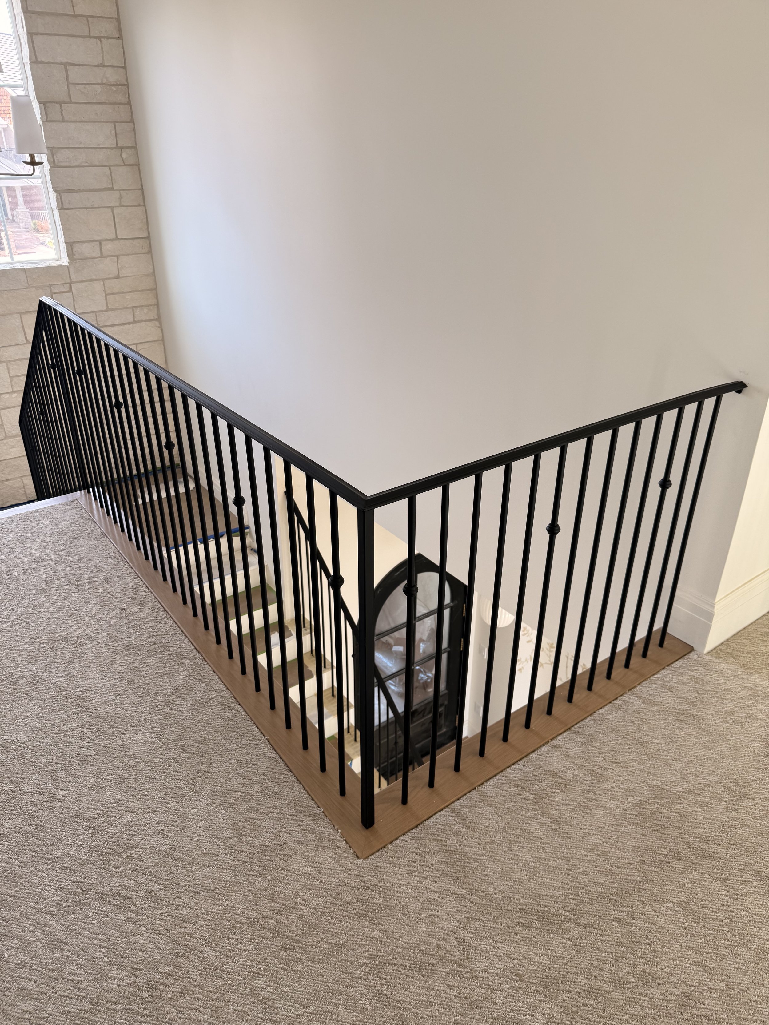 Image of a staircase with black metal railings, leading down to a lower level, with a white wall on the right and a brick wall near a window on the left.
