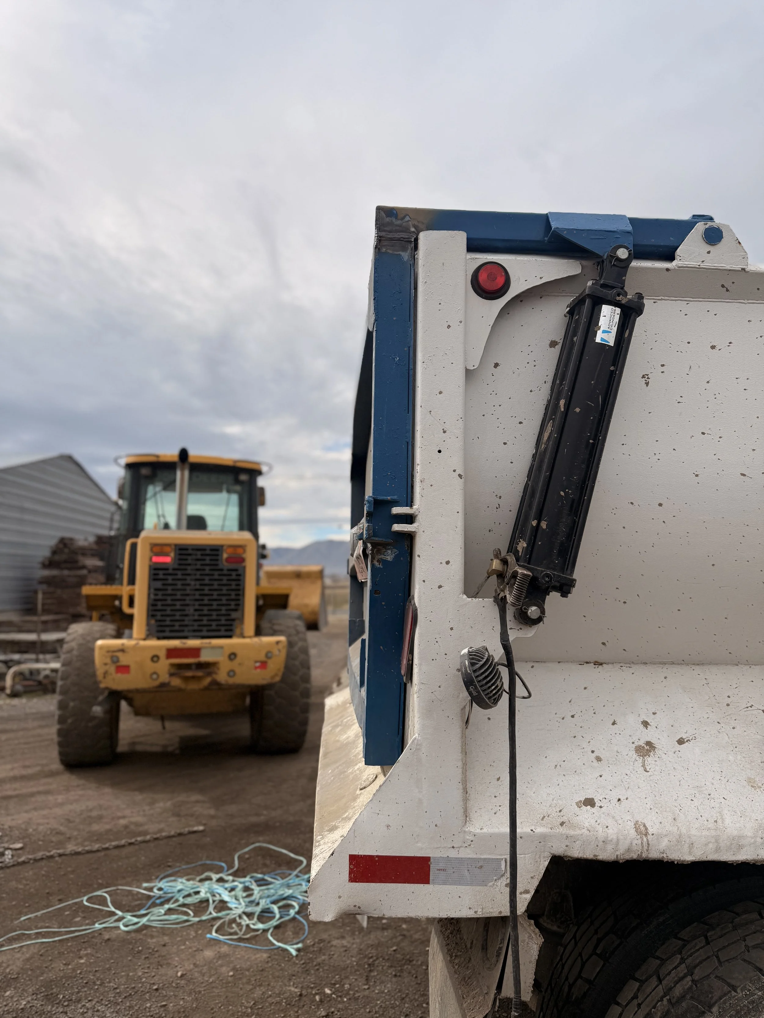 Close-up of the back corner of a white utility truck with a hydraulic lift, a tail light, and a tangled blue rope on the ground, with a large yellow construction vehicle and a gray building in the background under a cloudy sky.